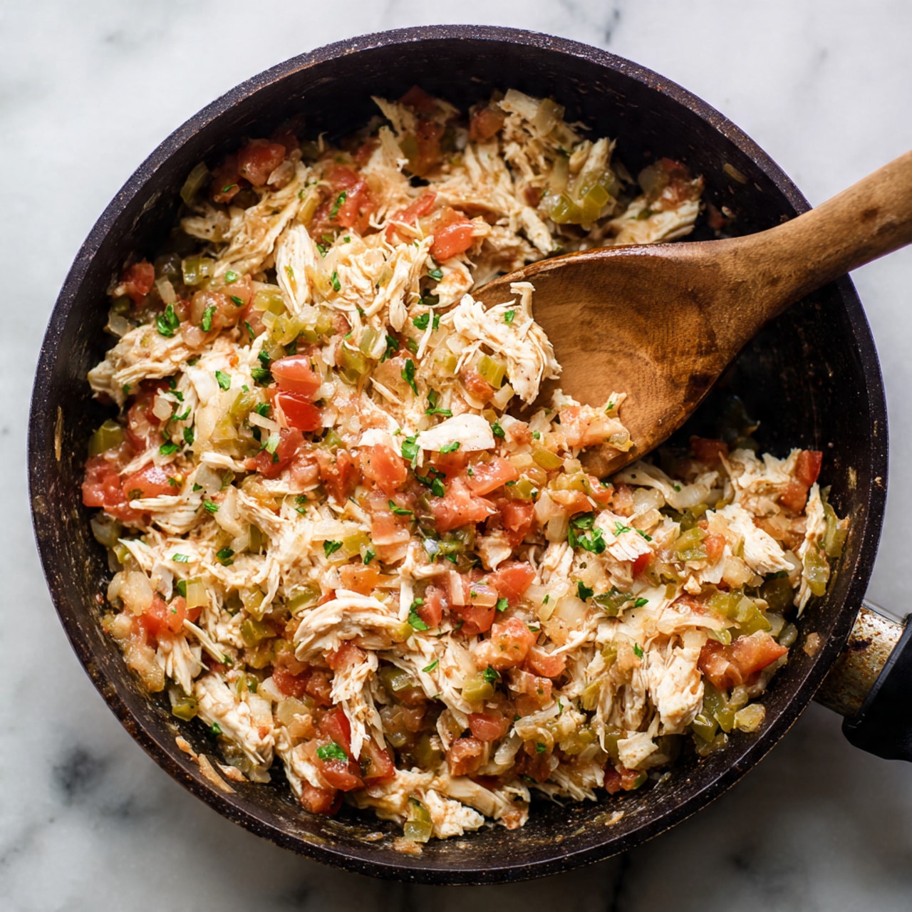 The image shows a close-up of a black pan filled with a mix of shredded white chicken, diced red tomatoes, light green pieces of cooked green chiles, and translucent cooked onions. The mixture looks soft and slightly chunky, with a rustic texture. A wooden spoon is scooping the mixture from the pan, showing the layers of the ingredients combined. The pan sits on a white marbled surface. photo taken with an iphone --ar 4:5 --v 7