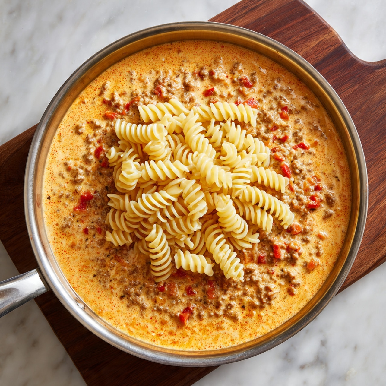 A close-up view of a pan filled with creamy pasta showing about three layers: spiral-shaped fusilli pasta in a pale yellow color mixed with a rich, light orange creamy sauce as the base layer; chunks of browned meat and diced red and yellow bell peppers spread evenly throughout; and small bits of green herbs sprinkled on top adding a fresh texture. A wooden spoon is resting in the pan, stirring the mixture. The pan sits on a wooden table next to fresh parsley, a whole onion, and a red bell pepper cut open, with a cookbook partially visible in the upper left corner. The background is a white marbled surface. Photo taken with an iphone --ar 4:5 --v 7
