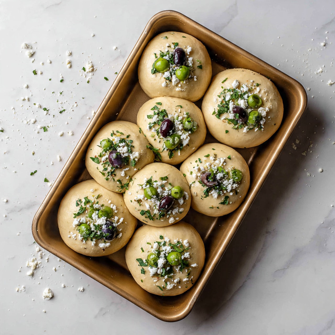 A golden brown rectangular baking tray filled with nine round, smooth dough balls arranged in three rows, topped with scattered white crumbled cheese and dried green herbs. Among the dough balls are a few pieces of green and dark purple olives peeking through, adding a pop of color. The tray sits on a white marbled surface, with some small crumbs around it. Photo taken with an iphone --ar 4:5 --v 7