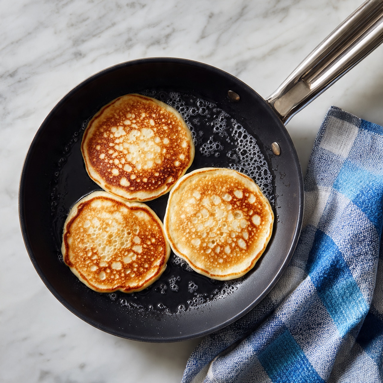 A black pan with three light golden pancakes cooking inside, each pancake round with small bubbles on the surface, showing a soft and slightly uneven texture. The pan sits on a white marbled surface with a blue and white checkered cloth partially visible on the right side. The metal handle of the pan extends to the left. Photo taken with an iphone --ar 4:5 --v 7