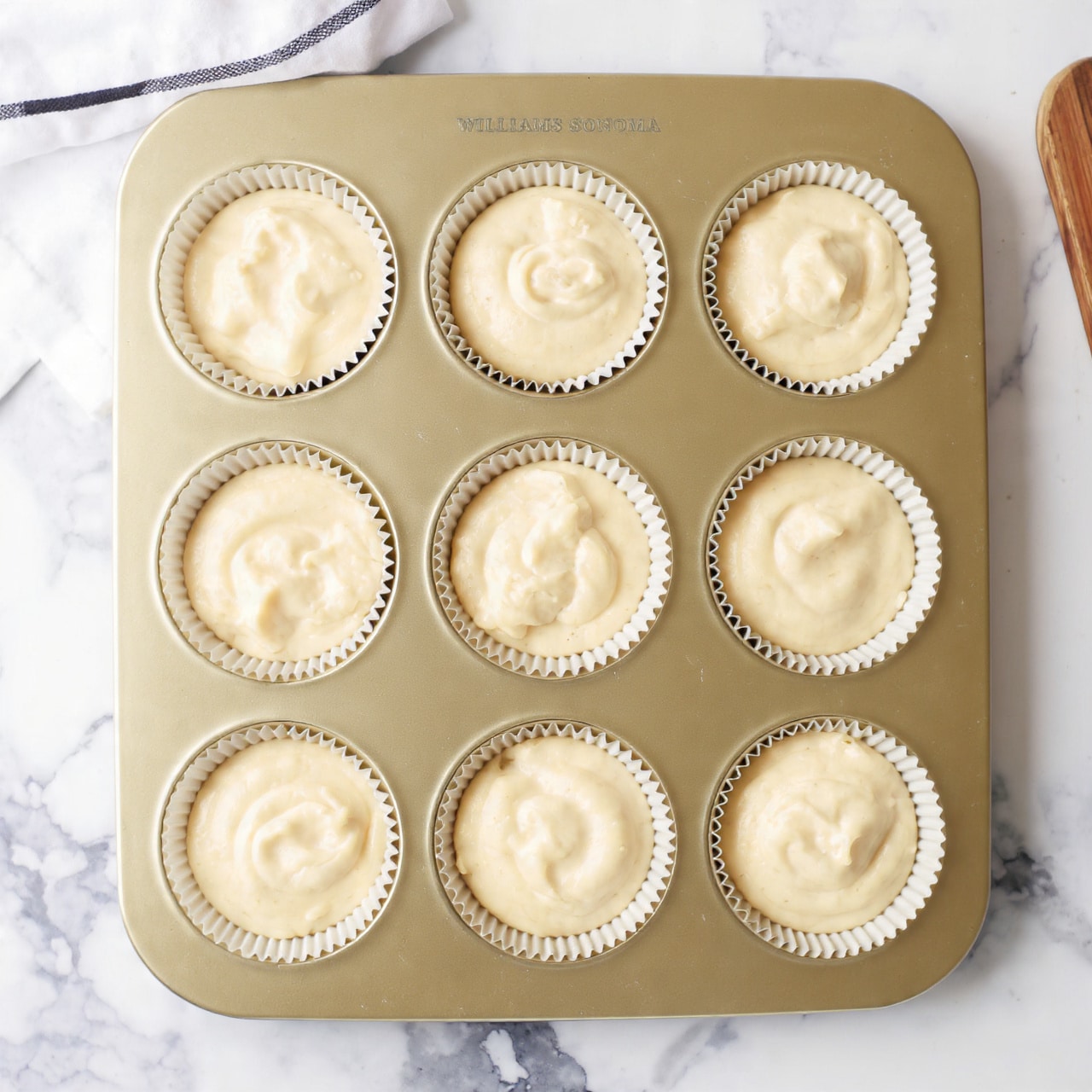 A golden muffin tray holds 12 round cups filled with light beige smooth batter, each slightly domed and showing small bubbles on the surface. The tray sits on a white marbled texture, giving a clean and bright background. The batter looks creamy and thick, evenly filling each cup almost to the top. The tray is a soft gold color with the