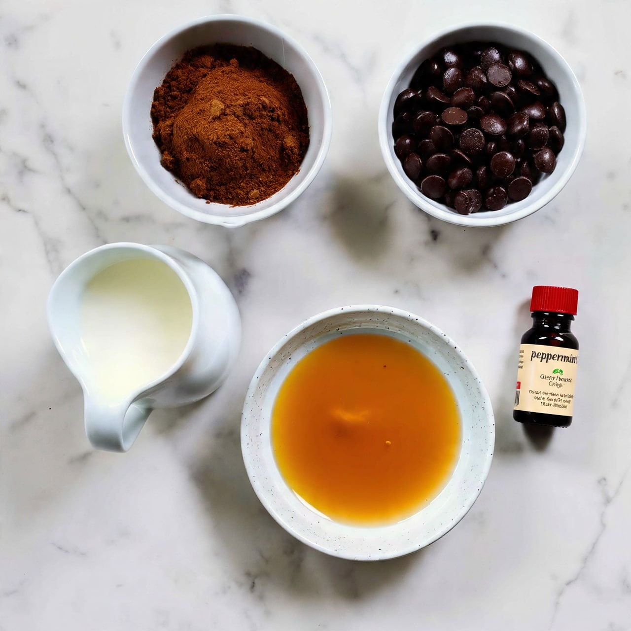 Four small white bowls with different ingredients and one small dark bottle are arranged on a white marbled texture. The top bowl contains dark brown cocoa powder with a soft, powdery texture. To the right, a bowl holds dark chocolate chips, small and shiny with a smooth surface. Below, a speckled white bowl is filled with golden maple syrup that looks thick and glossy. To the left, a white jug holds white dairy-free milk with a smooth liquid surface. Next to the syrup bowl is a small dark bottle with a red cap labeled peppermint extract. The items are spaced evenly, showing clear labels for each ingredient. Photo taken with an iphone --ar 4:5 --v 7