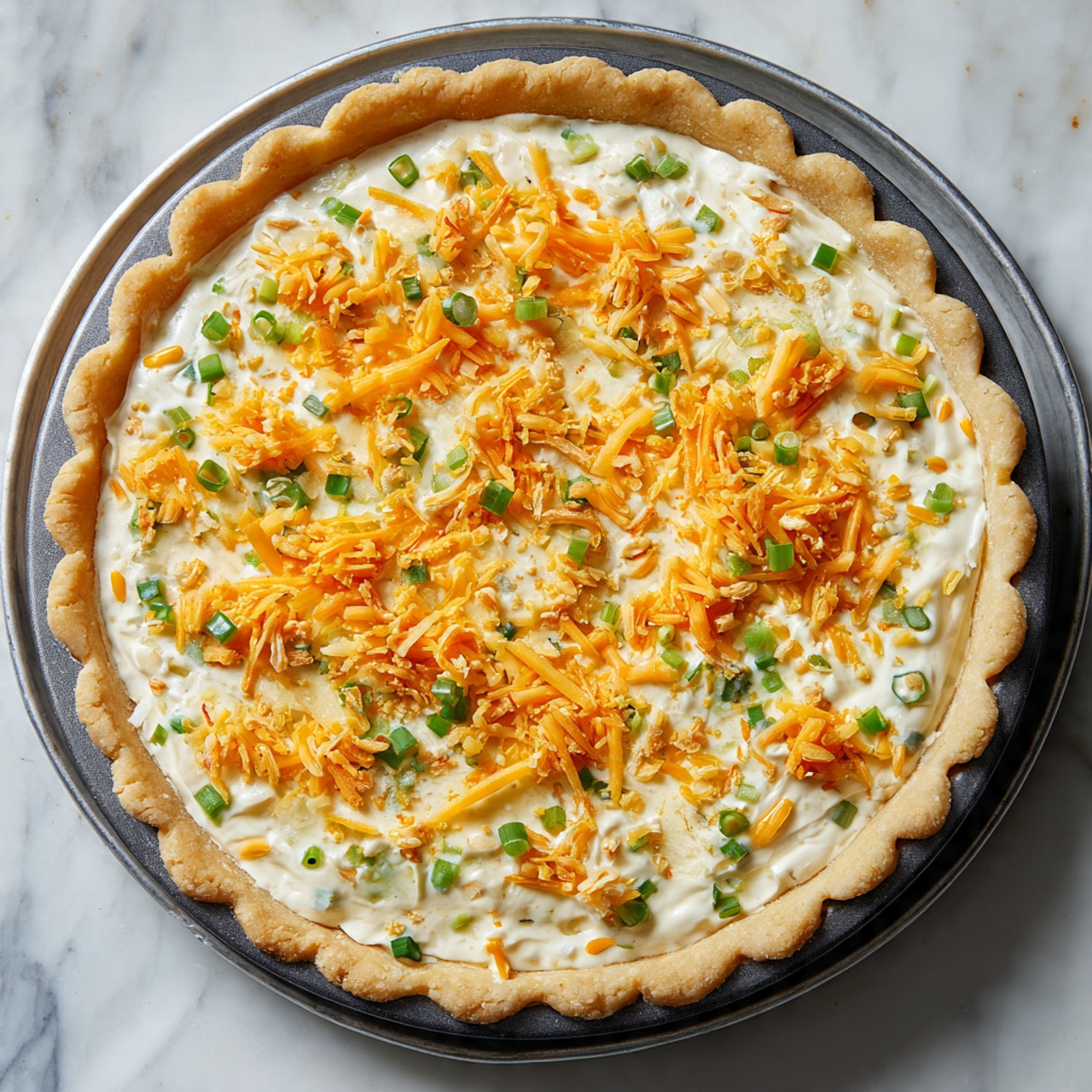 A round pie with a golden, scalloped crust forms the base layer. Inside the crust, there is a creamy, pale white mixture spread all over, filled with small green pieces of chopped scallions evenly scattered throughout. On top of the creamy layer, there are bright orange shredded cheese patches and lightly sprinkled shredded white cheese, creating a colorful contrast. The entire pie sits inside a dark round baking pan which rests on a silver metal tray. The background is a white marbled surface photo taken with an iphone --ar 4:5 --v 7