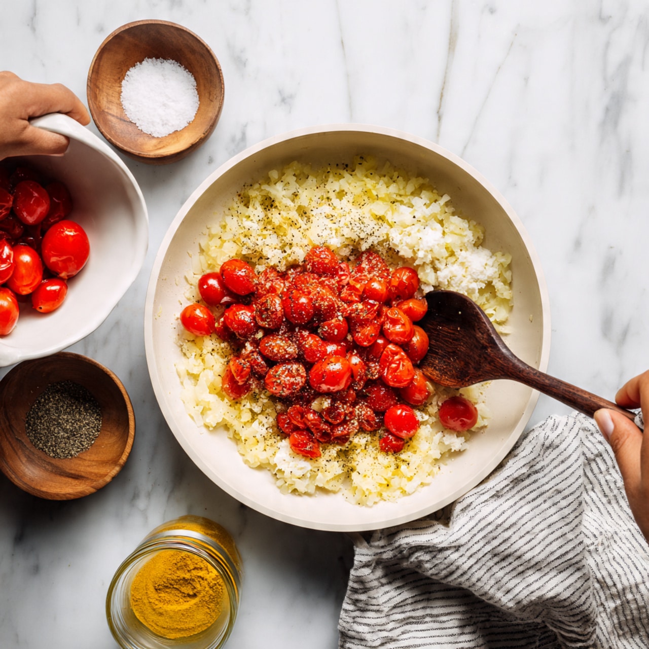 A white round pan sits on a white marbled surface with a striped cloth on one side. Inside the pan, there is a layer of cooked, chopped onions with a soft yellowish color and some black pepper seasoning scattered over. On top of the onions, vibrant red cherry tomatoes are being poured from a white bowl held by a woman's hand with a tattoo on the forearm. A dark wooden spoon rests in the pan among the onions. Near the pan on the surface are two small wooden bowls, one filled with coarse salt and the other with ground black pepper, as well as a small glass jar of yellow spice. Photo taken with an iphone --ar 4:5 --v 7