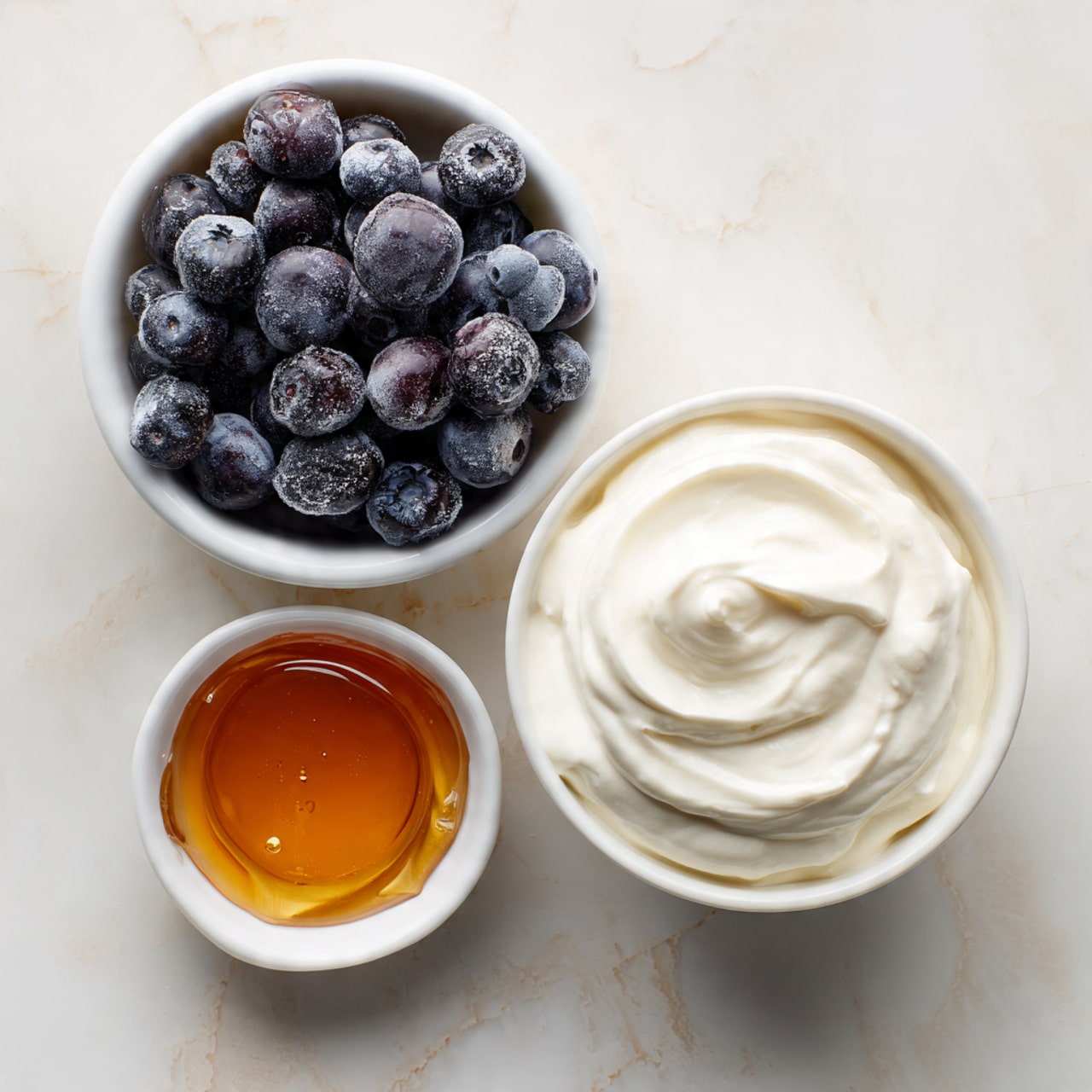 A top-down view shows three small white bowls placed on a white marbled surface. The largest bowl contains thick, creamy white Greek yogurt with smooth swirls on the surface. To the left, a smaller white bowl filled to the top with small, round frozen blueberries coated in frost appears dark purple and slightly dull due to the frost. Below them, a tiny white dish holds amber-colored maple syrup with a glossy, smooth surface. Each bowl is clearly labeled with simple black text: