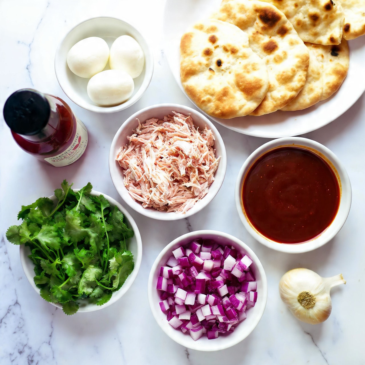 The image shows several small bowls and items arranged on a white marbled surface, each containing different ingredients for a dish. At the top right, there are three light brown mini naan breads stacked on a white plate, with slight browned spots. To the left, a small white bowl is filled with white mozzarella balls. Below that, another small white bowl holds green cilantro leaves with a rough texture. In the middle, a white bowl contains shredded light beige chicken. To the right of the chicken, a white bowl holds dark brown BBQ sauce with a smooth surface. Below the sauce, a white bowl is filled with purple and white chopped red onion. Next to the onion is a whole bulb of garlic with white outer skin. On the left side of the image is a dark brown bottle labeled