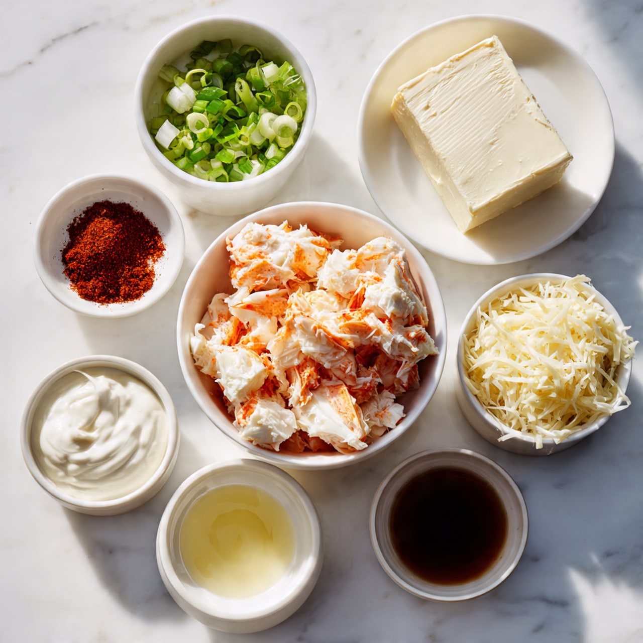 A white marbled surface holds seven white bowls arranged closely in a loose circle. At the center is a bowl filled with chunks of cooked crab meat with orange and white pieces. To the upper right, a white plate displays a block of smooth, cream-colored cream cheese. Surrounding the center bowl are smaller white bowls: top left with chopped green onions, middle left with a deep reddish-brown spice powder, lower left with thick white sour cream, lower right with shredded pale yellow cheese, middle right with a small amount of dark brown liquid, and bottom center with a clear watery liquid. The image has soft lighting and is cleanly arranged. Photo taken with an iphone --ar 4:5 --v 7