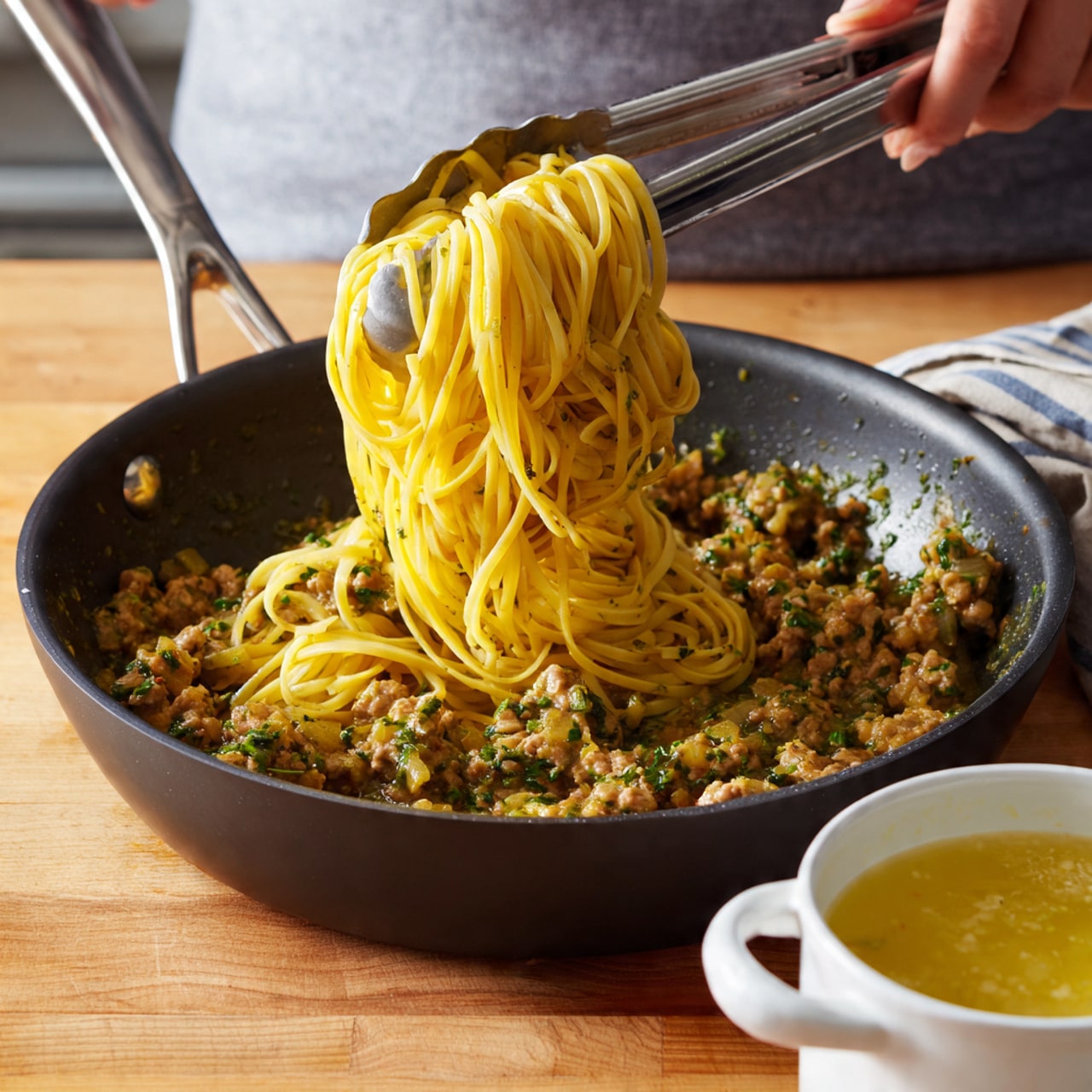 A close-up of a black cooking pan filled with greenish-yellow meat sauce mixed with small pieces of finely chopped ingredients. Over the pan, a woman's hand is holding silver tongs that lift a tight bundle of cooked yellow spaghetti pasta just above the sauce. The pan is on a wooden surface, and next to the pan on the right side is a small white pot with light yellow broth inside. Photo taken with an iphone --ar 4:5 --v 7