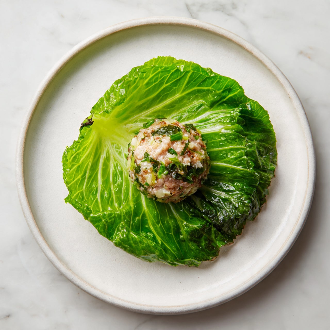A single large, bright green cabbage leaf is placed flat on a plain white plate, forming the bottom layer. On top, there is a round ball of raw minced meat mixed with visible small bits of white and green ingredients, centered on the cabbage leaf. The plate rests on a white marbled surface, giving a clean and simple look. photo taken with an iphone --ar 4:5 --v 7