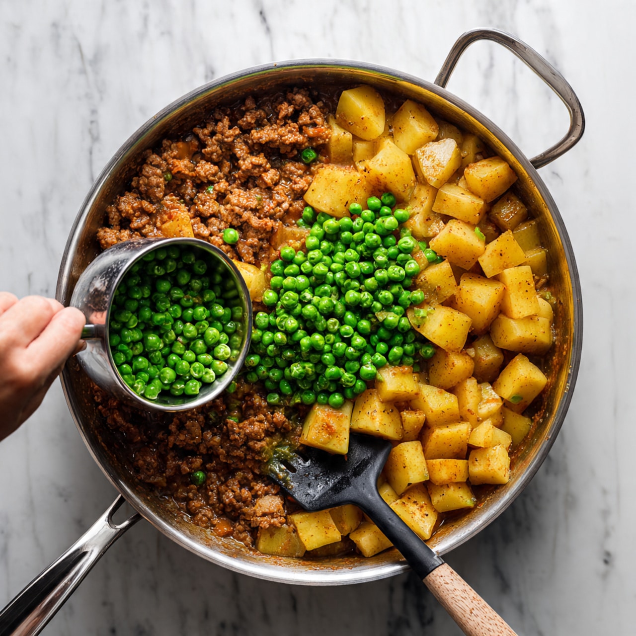 A silver pan filled with a mixed layer of cooked ground meat and cubed yellow potatoes, both coated in a brownish sauce, sits on a white marbled surface. Frozen green peas are being poured from a silver measuring cup held by a woman's hand over the meat and potatoes, adding a fresh green layer on top. A black spatula with a wooden handle rests inside the pan on the right side. photo taken with an iphone --ar 4:5 --v 7
