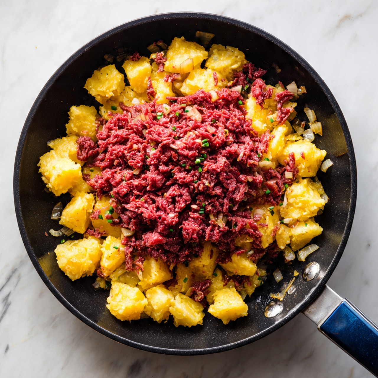 A black frying pan with a blue handle sits on a white marbled texture background. Inside the pan, there are two main layers: the bottom layer is golden yellow cooked diced potatoes with a slightly crispy texture, and on top is a generous pile of finely chopped pinkish-red corned beef. Small bits of sautéed onions are mixed into the potatoes. The colors show a contrast between the warm yellow of the potatoes and the rich red of the meat. The photo taken with an iphone --ar 4:5 --v 7
