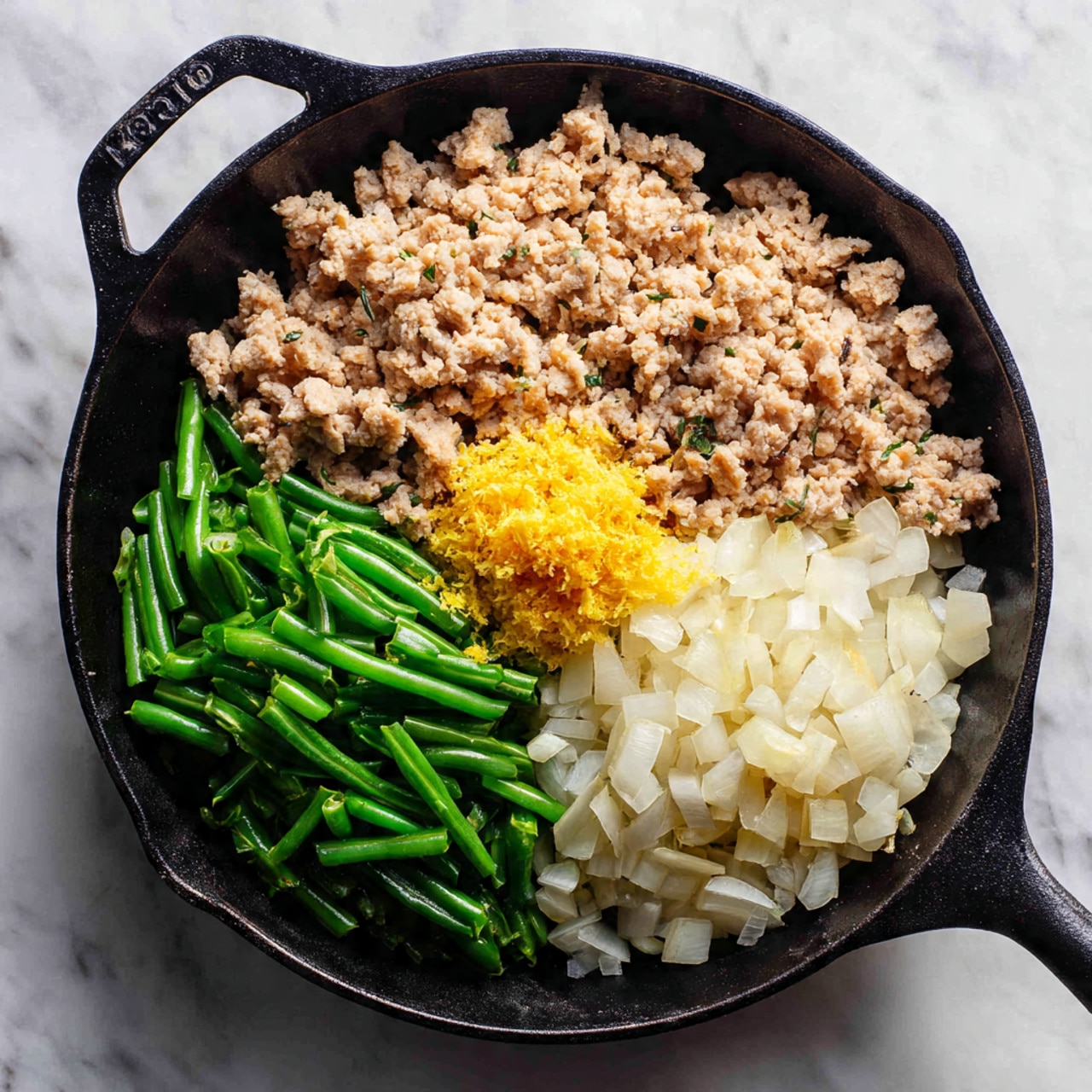 A black cast iron pan sits on a white marbled surface, filled with four distinct sections of ingredients. The largest section is filled with cooked ground meat that looks crumbly and light brown with small bits of onion mixed in. Below it is a mound of white chopped onions, diced into small pieces. To the left of the onions, there are fresh bright green sliced snap peas or green beans, cut diagonally. On top of the black pan’s surface, at the center between the meat and vegetables, is a small pile of finely grated yellow ginger. Photo taken with an iphone --ar 4:5 --v 7