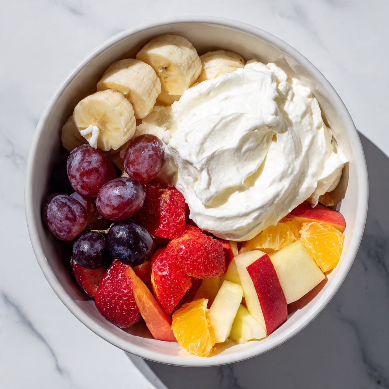 A large white bowl sits on a white marbled surface filled with a colorful mix of fresh fruit and a big dollop of thick white cream on one side. The fruit mix shows distinct layers including red and dark purple grapes, sliced light yellow bananas, bright orange segments, red strawberries cut in halves, and small pieces of yellow apples with red skin. The cream on the right side is smooth and fluffy, covering almost half of the fruit. The bowl shows a casual, fresh mixture ready to be stirred together, with sunlight casting soft shadows around the fruit and cream. photo taken with an iphone --ar 4:5 --v 7