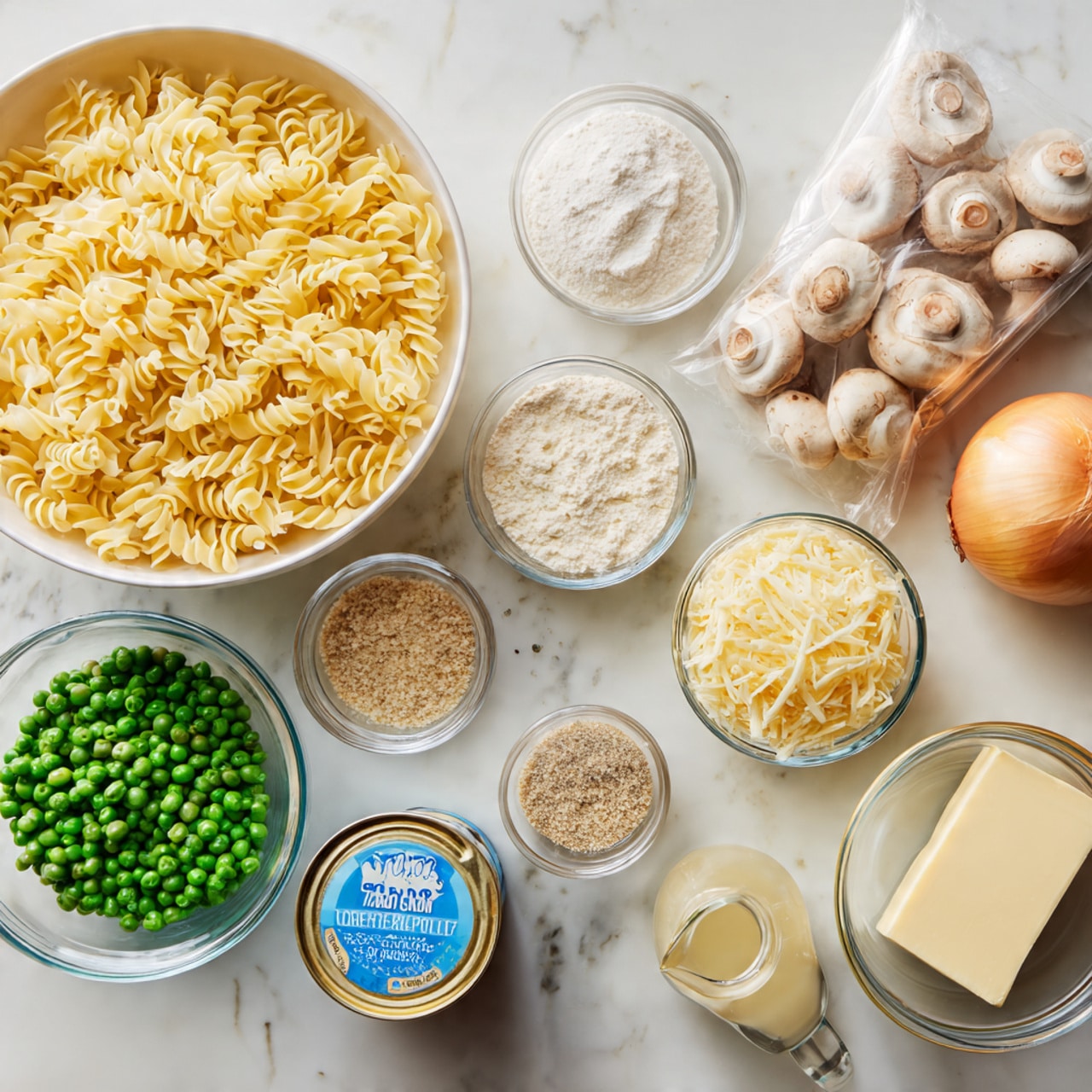 In a silver pan on a white marbled surface, there are three main sections filled with different foods. On the right side, there is a large portion of cooked, yellow curly egg noodles with a soft texture. On the left side, there are pieces of light brown tuna that look soft and flaky. Beneath the tuna and spread throughout the left side is a creamy white sauce with small green peas visible inside. The pan has a shiny, silver handle that points to the left side of the image. Photo taken with an iphone --ar 4:5 --v 7