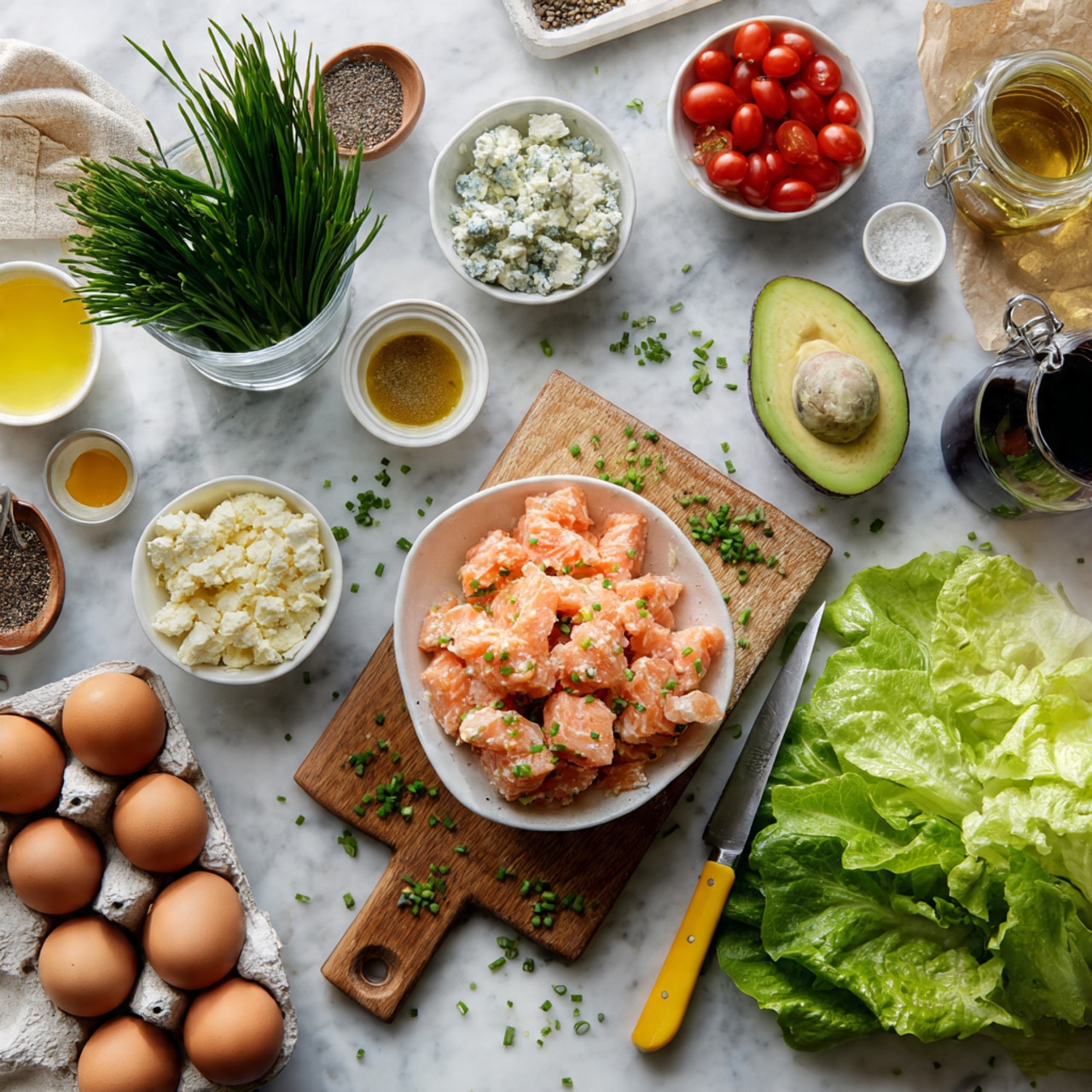 The image shows fresh ingredients arranged on a white marbled surface, centered around a wooden cutting board. On the board is a white bowl filled with pinkish-orange cooked salmon chunks, scattered chopped chives next to a knife with a yellow handle, and green leafy lettuce leaves on the side. Surrounding the board are other small white bowls and measuring cups containing red cherry tomatoes, crumbled white and blue cheese, cooked crispy bacon pieces, and a small amount of mustard. There is a glass cup filled with fresh green chives at the top left, and nearby are three brown eggs in a brown cardboard carton, an avocado, and a tiny white bowl with honey. A clear jar with black pepper and salt sits near the top right, along with measuring cups of dark red wine vinegar and golden olive oil. Everything is neatly placed with bright natural light, photo taken with an iphone --ar 4:5 --v 7