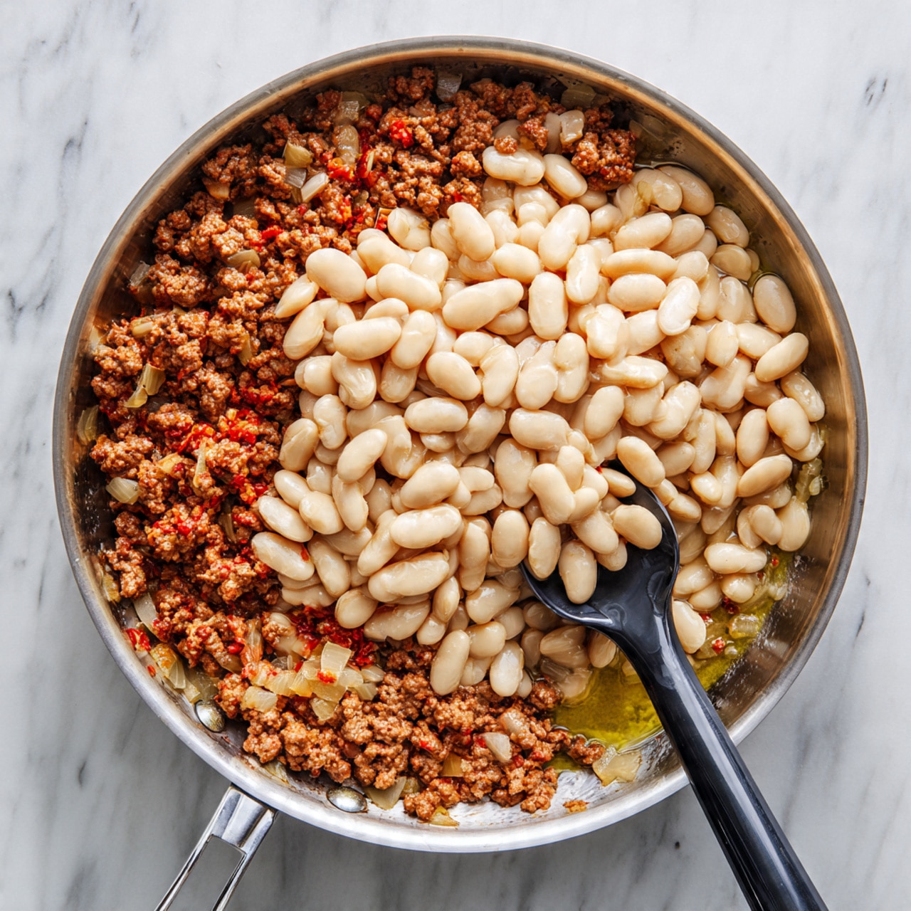 A silver metal pan sits on a white marbled surface, filled with two main layers. The bottom layer is made of cooked ground meat mixed with small pieces of diced onions, showing a rich brown color with some red oil around the edges. On top of this, there is a large pile of creamy white beans that cover the center of the meat. A black and silver spoon rests inside the pan, partially mixing the meat layer, with the spoon handle extending out over the pan edge. Photo taken with an iphone --ar 4:5 --v 7