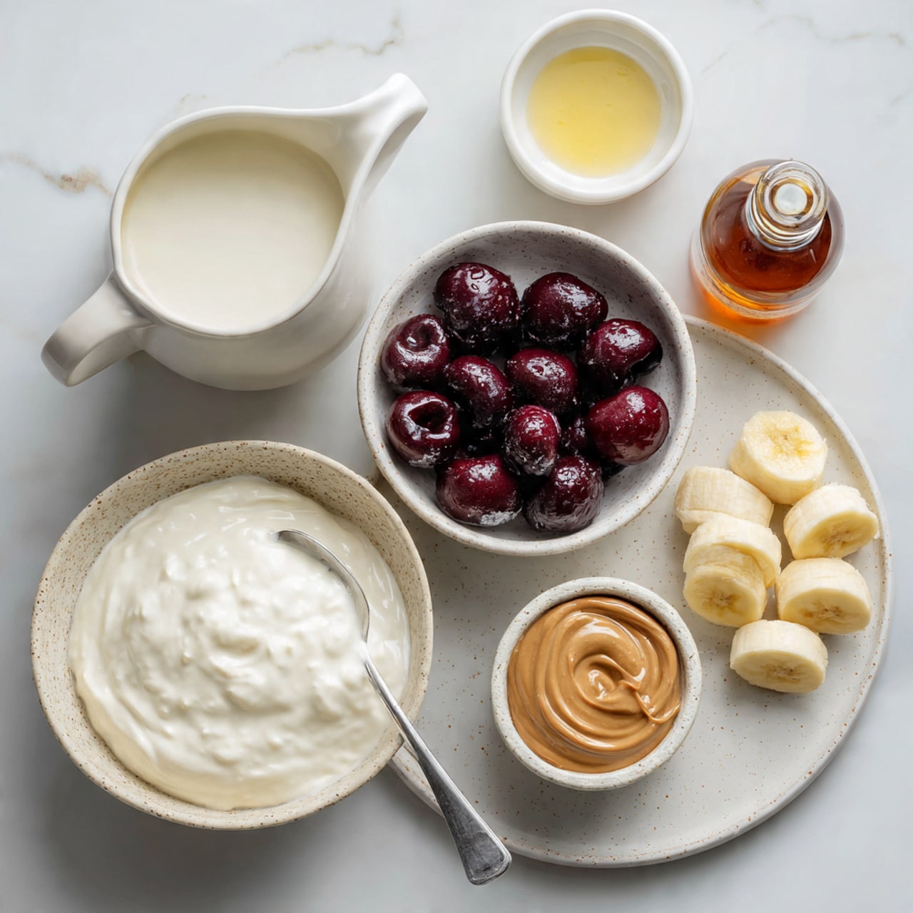 The image shows a white marbled surface holding six items: a white ceramic jug filled with milk on the top left, a small white bowl with a yellowish liquid residue to the right of the jug, and an amber bottle of almond extract to the far right. Below these, there is a white speckled bowl filled with dark red frozen cherries with visible frost on them, a white speckled bowl containing thick white yogurt with a spoon inside on the left side, and a white speckled plate at the bottom holding two peeled banana halves. To the right of the plate is a small white speckled bowl with a swirl of brown almond butter. Photo taken with an iphone --ar 4:5 --v 7