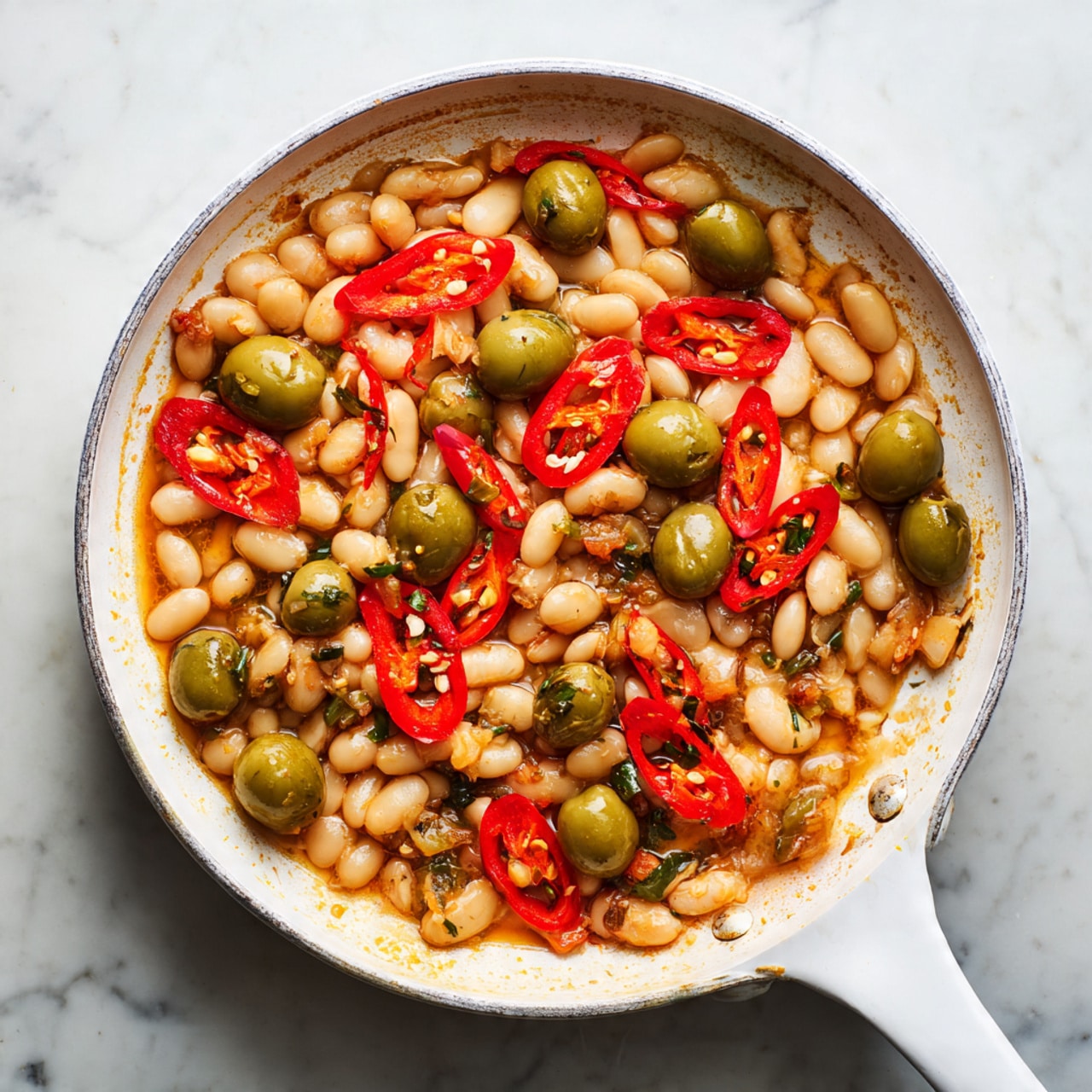 This image shows a white pan with a long handle placed on a white marbled surface. Inside the pan, there is one layer of cooked white beans scattered evenly across the bottom. Mixed in with the beans are whole and halved green olives, some with a smooth texture and a bright green color. Thin slices of red chili pepper are spread throughout, adding bright red tones and a slight shine. The pan interior is slightly stained with light brown cooked bits. Photo taken with an iphone --ar 4:5 --v 7