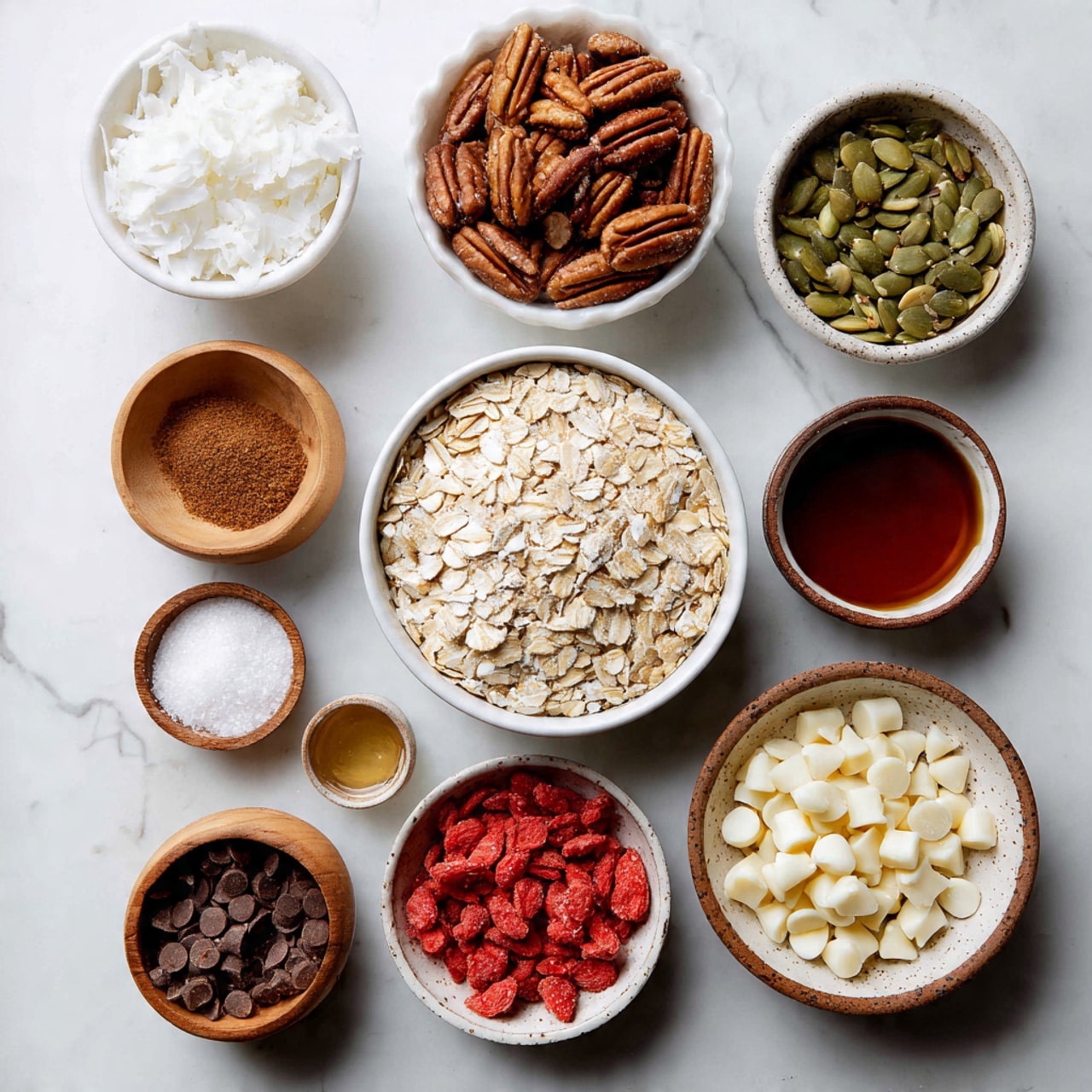 Several small bowls and cups arranged neatly on a white marbled surface hold different ingredients for a recipe. In the center is a white bowl filled with light beige rolled oats, surrounded by a white bowl on the left with large white coconut flakes. Near it is a small wooden bowl with white coarse salt and a tiny wooden bowl of cinnamon powder below it. To the top right of the oats is a white bowl with chopped pecans, a white bowl with green pumpkin seeds, and a white bowl with chopped almonds. On the right side, a white bowl with speckled brown edges holds bright red freeze-dried strawberry pieces. Below the oats is a white bowl full of small white chocolate chips. Three small cups containing clear liquid, dark brown syrup, and vanilla extract, are placed among the bowls. The whole setup is bright, with a clean and natural look. photo taken with an iphone --ar 4:5 --v 7
