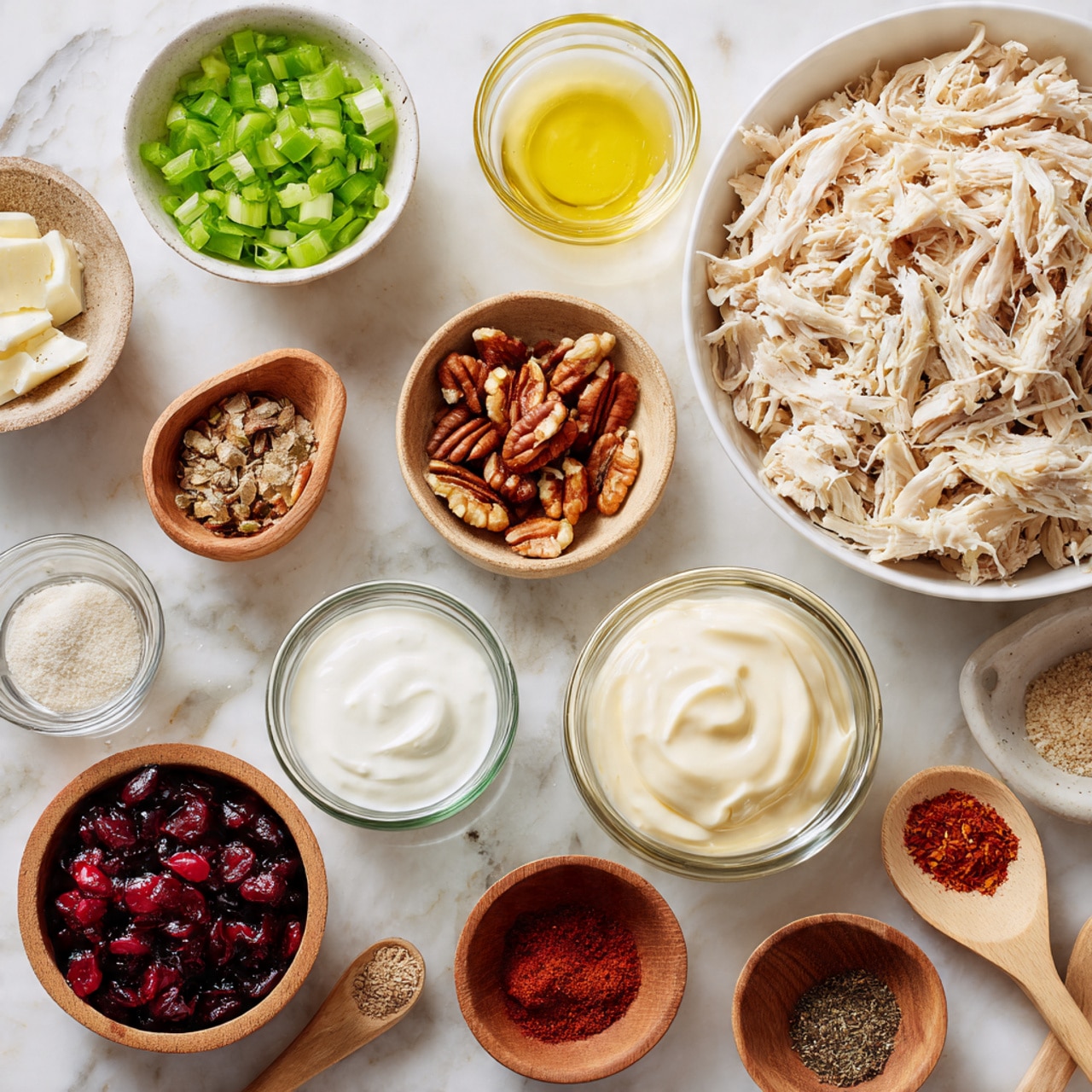 The image shows a top-down view of several small white bowls and glass containers arranged on a white marbled surface, each holding a different ingredient. At the top right is a large white bowl full of shredded chicken, light beige in color with a fibrous texture. Moving around counterclockwise, there is a small white bowl with bright green chopped celery, next to it a small white bowl with finely chopped sweet onion, and a small white bowl with mixed brown pecans. Near the center are small glass containers holding creamy white mayo, thick white Greek yogurt, and pale yellow honey. Also visible are a small white bowl with light beige garlic powder, a wooden bowl with vibrant red paprika, a rustic bowl with coarse black pepper, another wooden bowl with coarse white salt and a wooden spoon resting inside it, and a medium white bowl with rich red dried cranberries. A very small glass container holds light yellow lemon juice. Each ingredient is clearly labeled with text, and the whole setting is clean and organized. Photo taken with an iphone --ar 4:5 --v 7