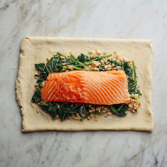 Two golden brown pastries sit side by side on a metal cooling rack over a white marbled surface. Each pastry has a top layer of puff pastry with a braided pattern of diagonal cuts that reveal bits of green leafy filling inside. The pastries are rectangular with slightly rounded edges, and the crust has a shiny, crisp texture from a baked glaze. The background is softly blurred, focusing on the warm, flaky pastries. Photo taken with an iphone --ar 4:5 --v 7