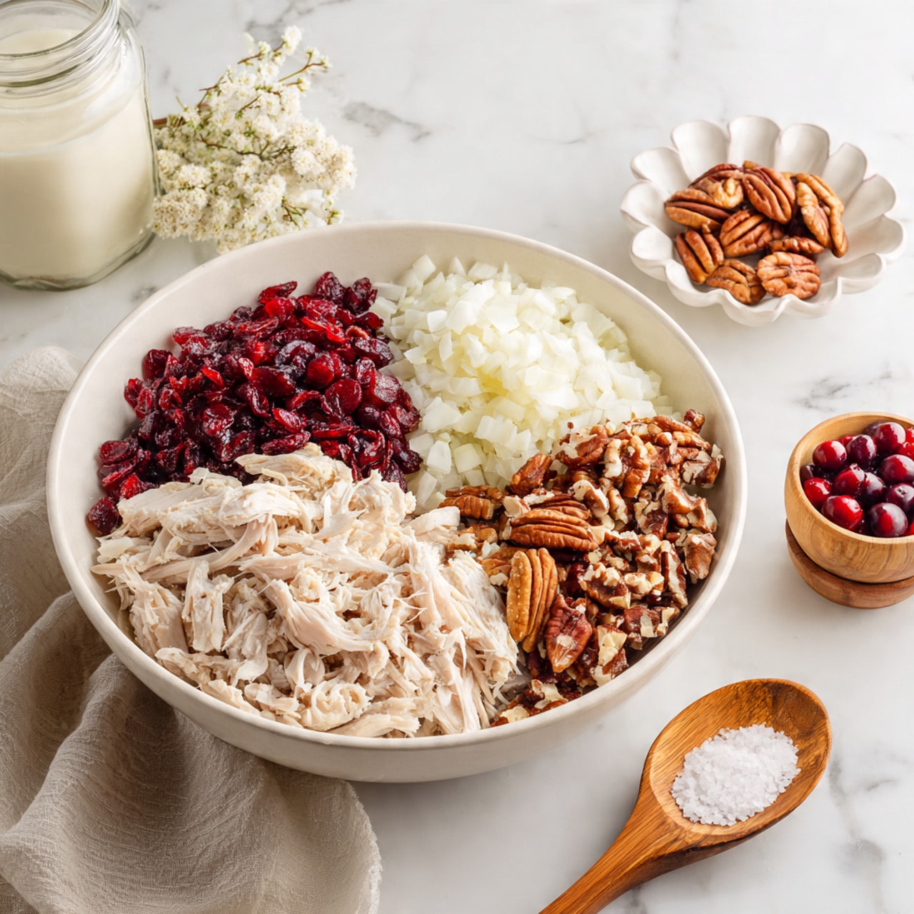 A large white bowl sits on a white marbled surface, filled with four distinct layers of ingredients arranged side by side. The first layer is shredded pale chicken with a soft texture. Next to it is a pile of small, white chopped onions, followed by a vibrant cluster of red dried cranberries. Adjacent to the cranberries is a section of chopped brown pecans with a rough texture. In front of the bowl, a small white dish holds additional dried cranberries, while above, a small white flower-shaped dish contains more pecans. To the left, there is a clear glass jar with a white creamy ingredient, and to the right, a small wooden bowl filled with coarse salt and a wooden spoon completes the scene. The photo taken with an iphone --ar 4:5 --v 7