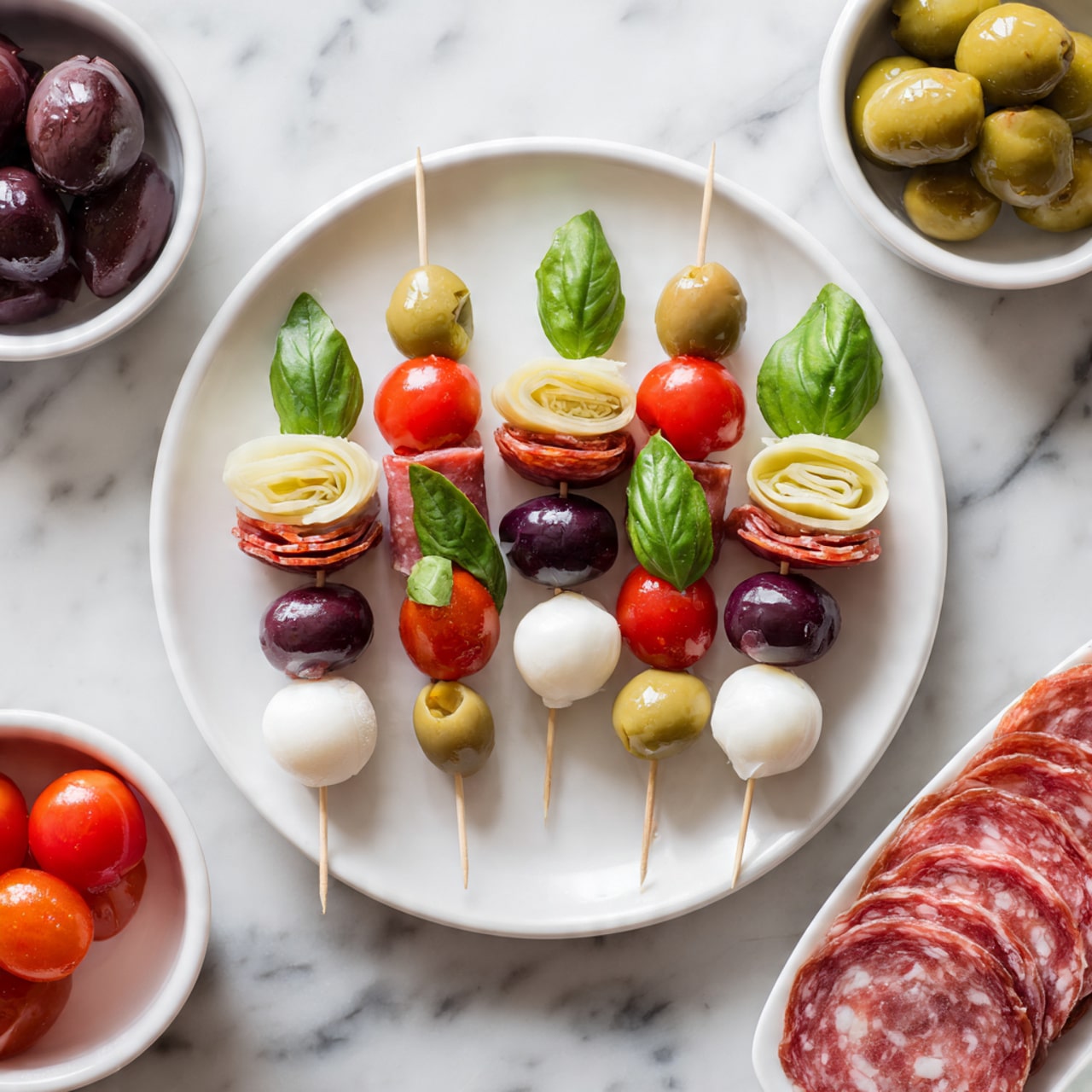 The image shows small skewers arranged on a large white plate with layers that include a dark green olive, a slice of pale artichoke, folded slices of reddish-pink salami, a bright red grape tomato, a fresh green basil leaf, and a white mozzarella ball. Around the large plate, there are small white bowls with dark purple olives, red grape tomatoes, white mozzarella balls, fresh green basil leaves, green olives, and chopped pale artichoke hearts. There is also a white plate at the bottom left corner with overlapping slices of reddish-pink salami. The background surface is a white marbled texture. photo taken with an iphone --ar 4:5 --v 7