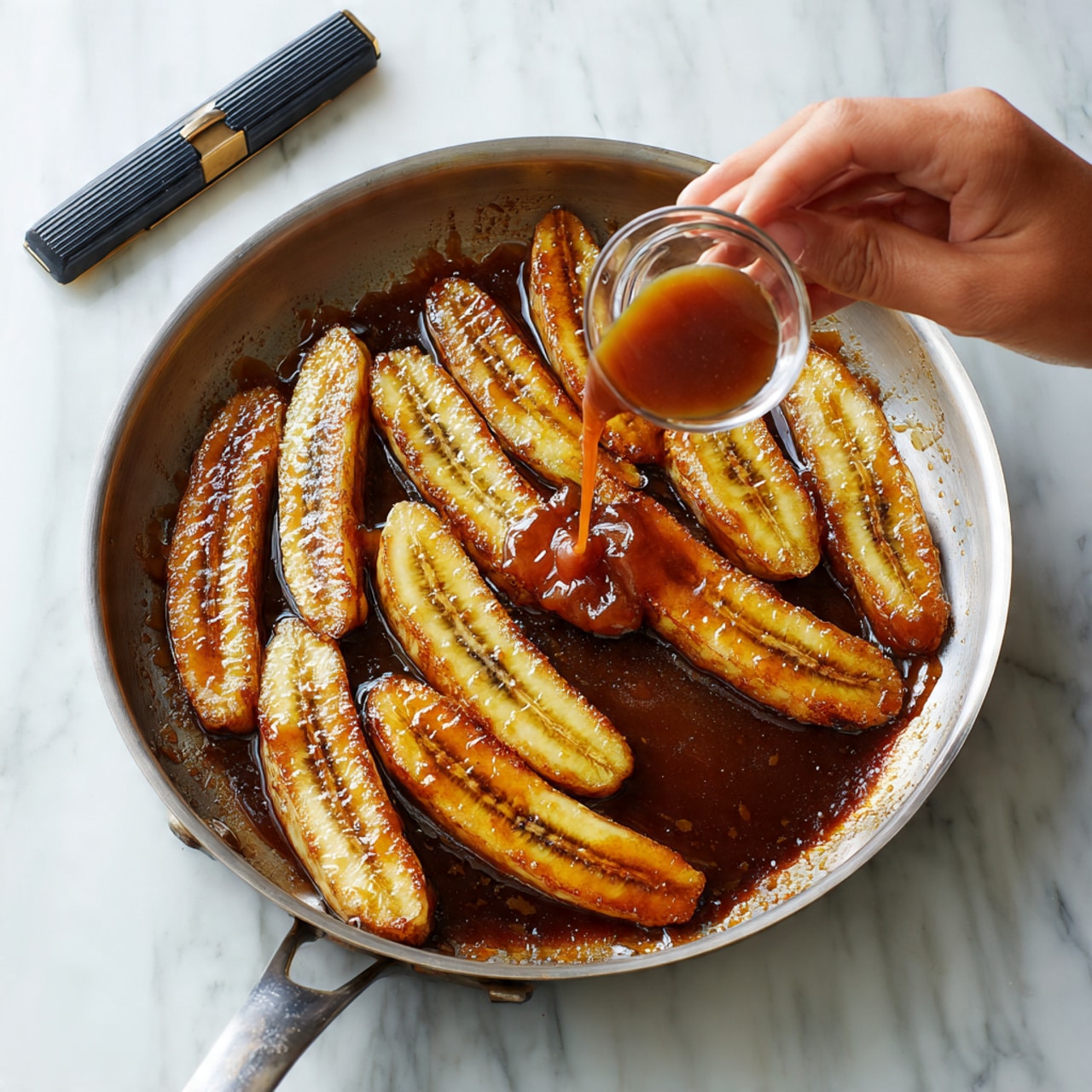 A silver pan is shown on a white marbled surface, with around eleven golden brown slices of plantains lying flat in dark, bubbling caramel sauce. A woman's hand is pouring more caramel sauce from a small clear glass onto the right side of the pan. The sauce adds a shiny glaze to the plantains, which have a slightly soft texture with light ridges from slicing. A black and gray lighter rests above the pan. photo taken with an iphone --ar 4:5 --v 7