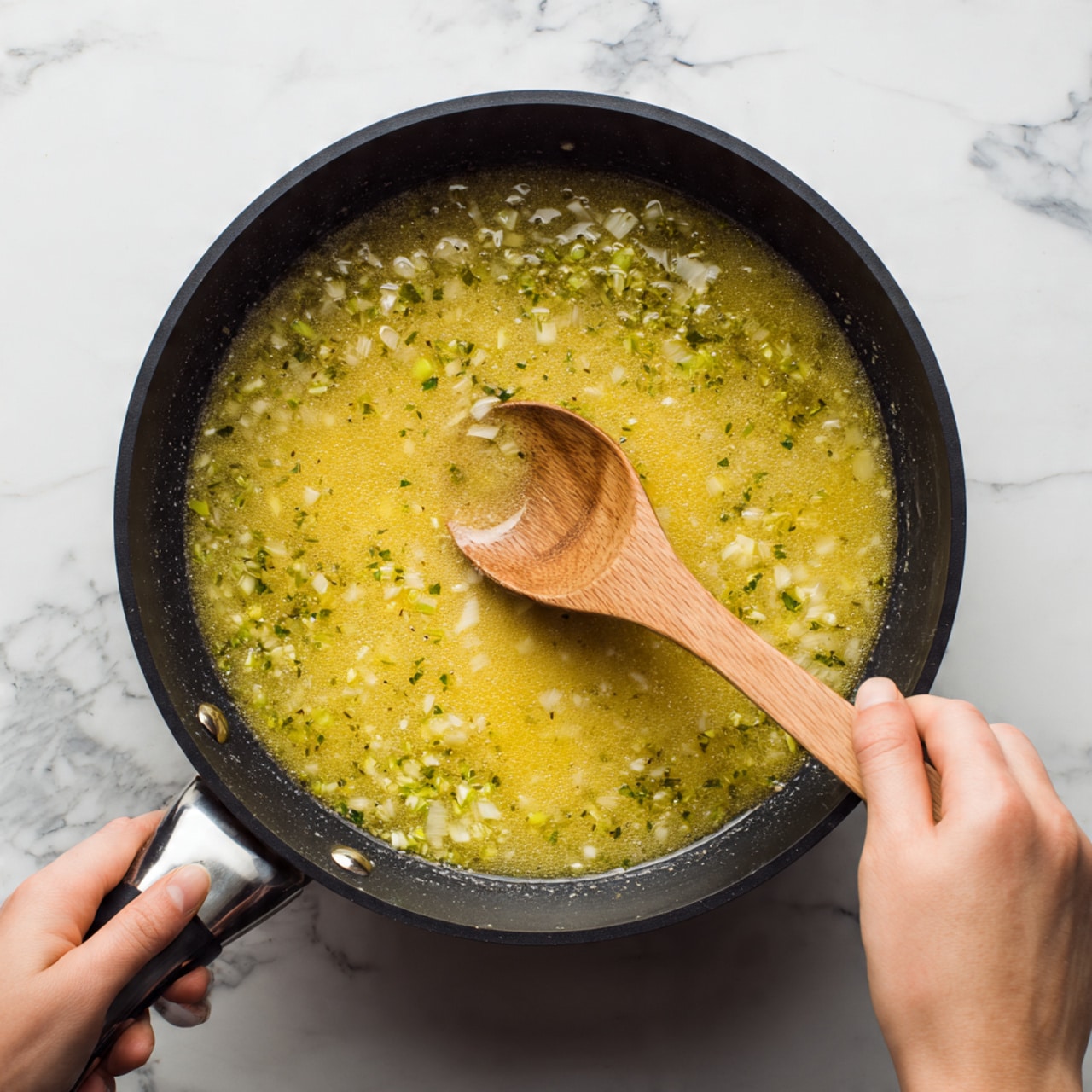The image shows a top view of a black frying pan filled with a yellowish sauce mixed with small greenish pieces of food, likely herbs or vegetables, and small sliced ingredients cooking together. A woman's hand holding a wooden spoon stirs the mixture from the right side, with the spoon partially submerged in the sauce. The pan is placed on a white marbled surface that contrasts with the pan and sauce colors. Photo taken with an iphone --ar 4:5 --v 7