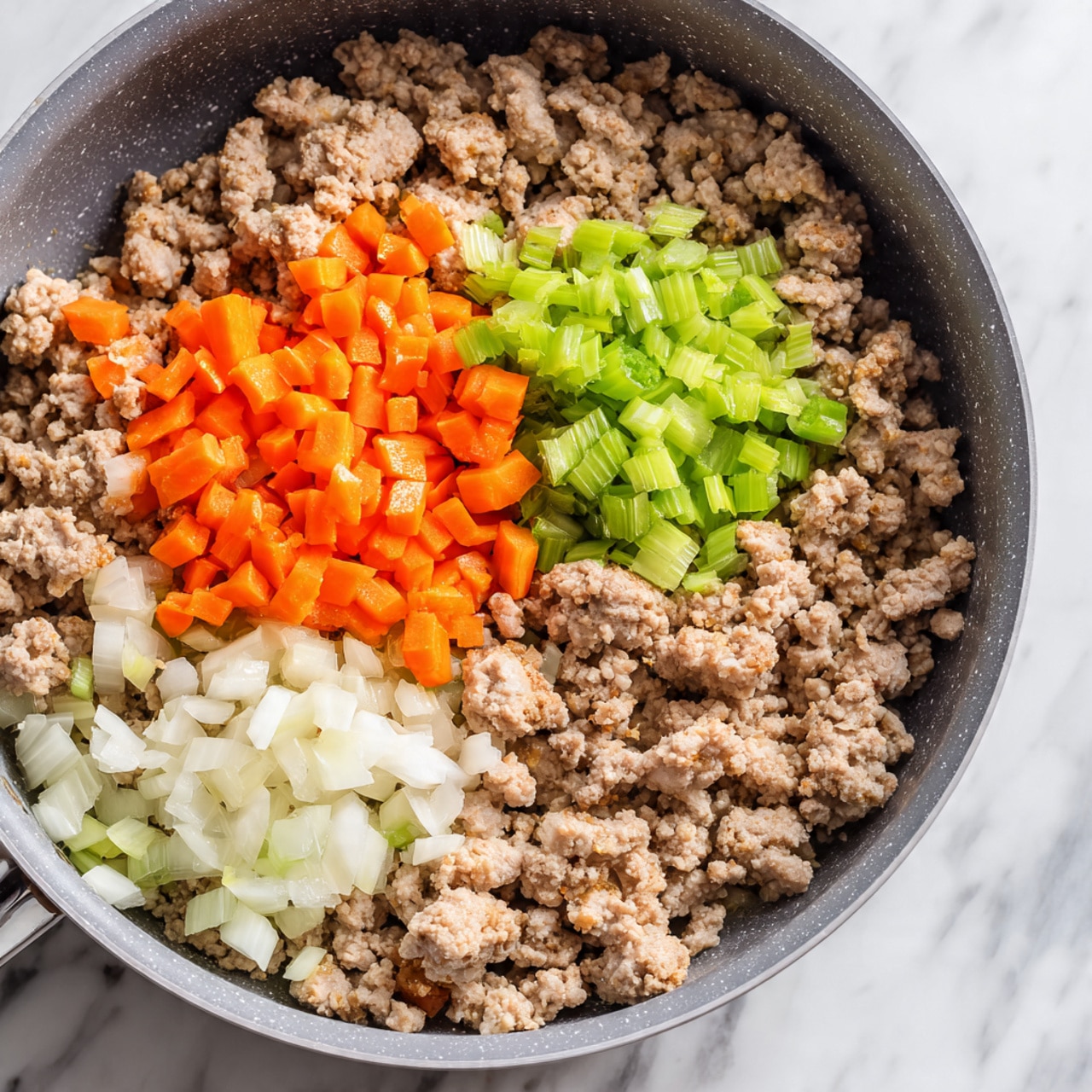 A close-up top view of a grey pan filled mostly with cooked ground meat that is light brown and crumbly in texture. On top of the meat, there are three distinct small piles of chopped vegetables: bright orange carrots, light green celery, and white onions with some small pieces of orange carrot mixed in. The pan sits on a white marbled surface. photo taken with an iphone --ar 4:5 --v 7
