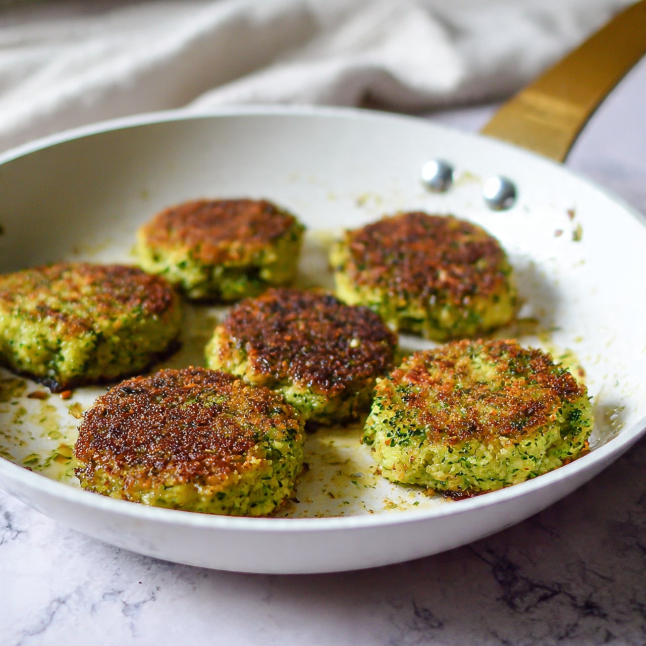 In the white frying pan with a gold handle, there are four small patties made mostly of green broccoli pieces mixed with bits of light beige grains or seeds. The patties have a rough, uneven texture with some parts browned and crispy, showing a toasted light brown color. They sit spaced apart on the bottom of the pan, which has a few small brown spots and a shiny layer of cooking oil. The background under the pan is a white marbled texture. photo taken with an iphone --ar 4:5 --v 7