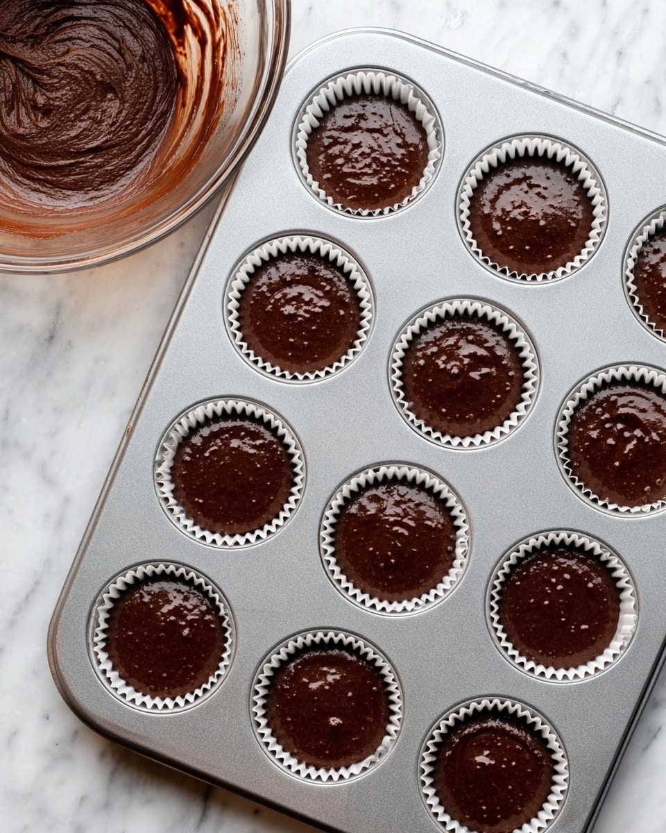 A 12-slot silver metal muffin tray sits on a white marbled surface, each slot lined with white ridged paper liners filled with shiny, rich dark brown chocolate batter. The batter fills almost to the top of each liner, showing a thick and smooth texture with slight swirls and small bubbles. On the left side of the image, part of a clear glass bowl with some chocolate batter inside is visible, resting also on the white marbled surface. The whole scene is bright and clean, focusing on the ready-to-bake chocolate cupcakes. photo taken with an iphone --ar 4:5 --v 7