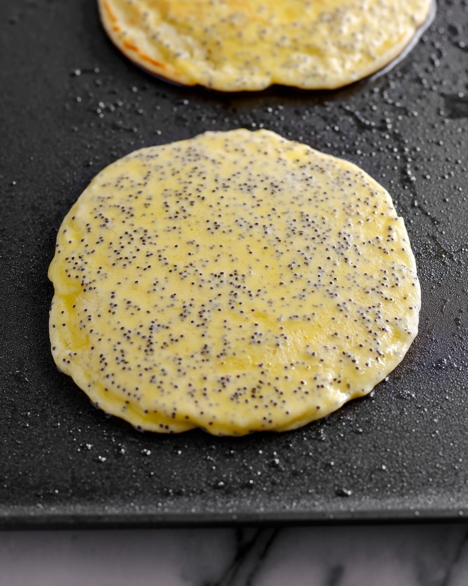 A close-up of a single pancake cooking on a dark non-stick griddle shows a light yellow batter speckled with many small black poppy seeds. The pancake has a soft and slightly bubbly texture with uneven edges, and a second pancake is partially visible above it on the griddle. The griddle surface glistens with small droplets of oil, and the background beneath the griddle is a white marbled texture. Photo taken with an iphone --ar 4:5 --v 7