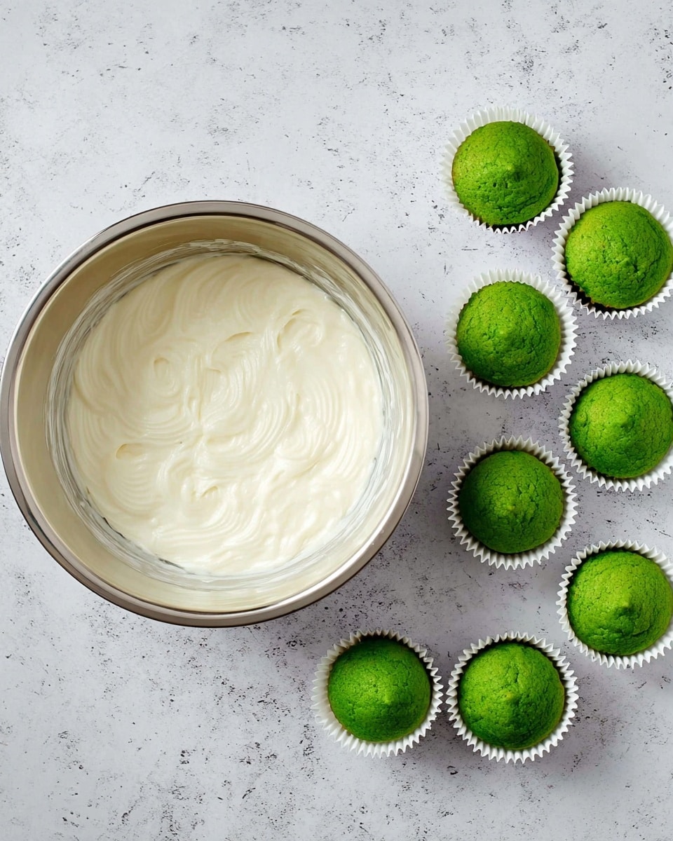 The image shows a metal mixing bowl filled with smooth, creamy white frosting with soft swirls across the surface, placed on a white marbled texture. To the right of the bowl, there are nine small white paper cupcake liners arranged in three rows of three, each holding a vibrant green cupcake with a slightly domed top and a soft texture visible on the surface. The cupcakes’ green color contrasts vividly with the white liners, and the overall scene looks clean and neatly organized. photo taken with an iphone --ar 4:5 --v 7