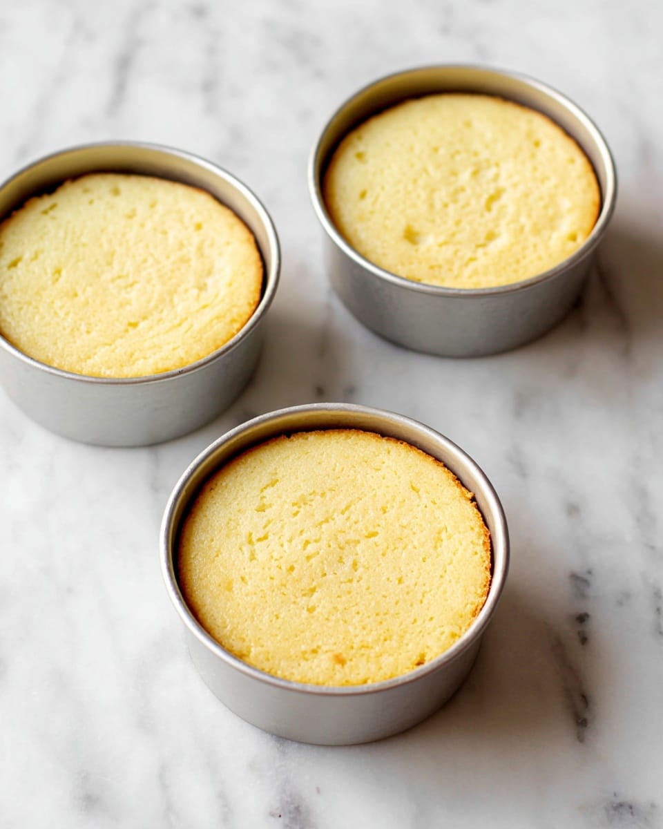 The image shows three small round cakes fresh out of the oven, each in a silver baking pan. The cakes are light yellow with a soft texture and slightly golden edges. They sit on a white marbled surface, evenly spaced in a triangular layout. The cakes are smooth on top with no decorations or toppings visible, and their height suggests they are fluffy and light. The silver pans have a shiny, clean look, contrasting with the soft, warm color of the cakes inside. photo taken with an iphone --ar 4:5 --v 7