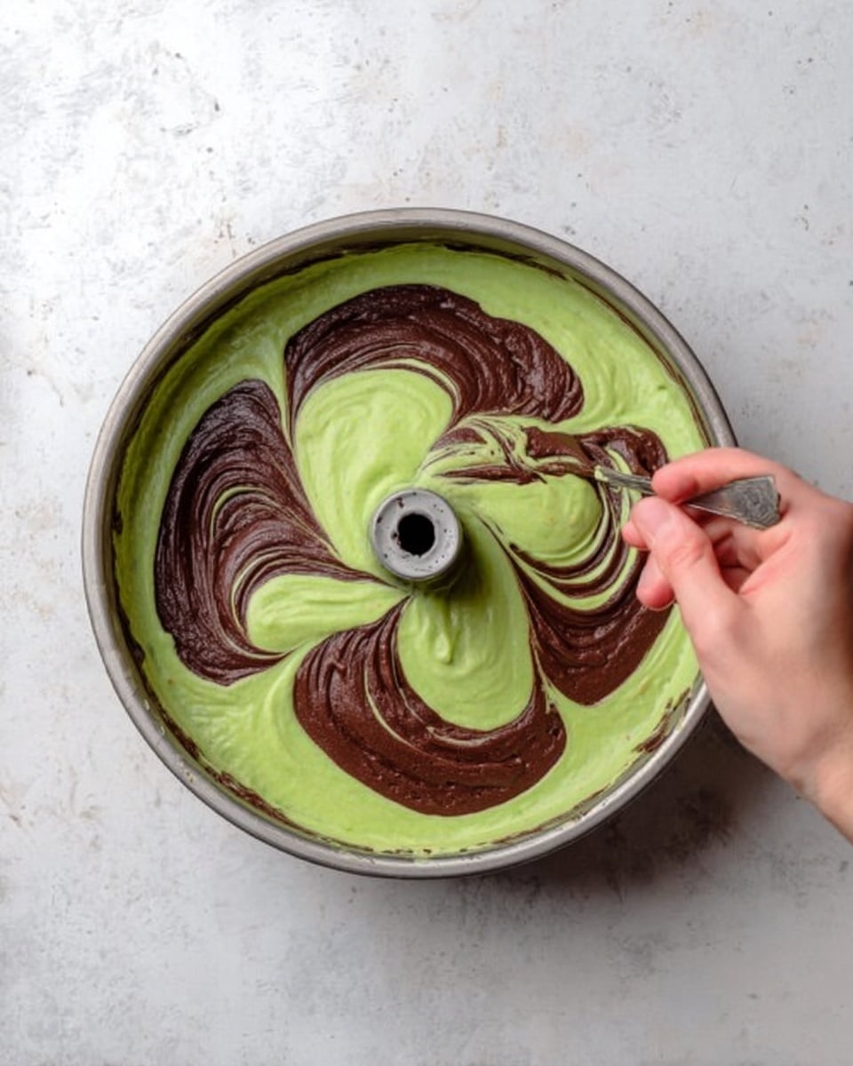 A silver bundt cake pan is filled with green batter as the base layer, smooth and creamy in texture. On top, swirls of thick dark brown batter spread artistically in a circular pattern across the green batter. A woman's hand holding a small spoon is gently dragging through the batters to create a marbled effect, with parts of both colors blending. The cake pan sits on a white marbled surface. photo taken with an iphone --ar 4:5 --v 7