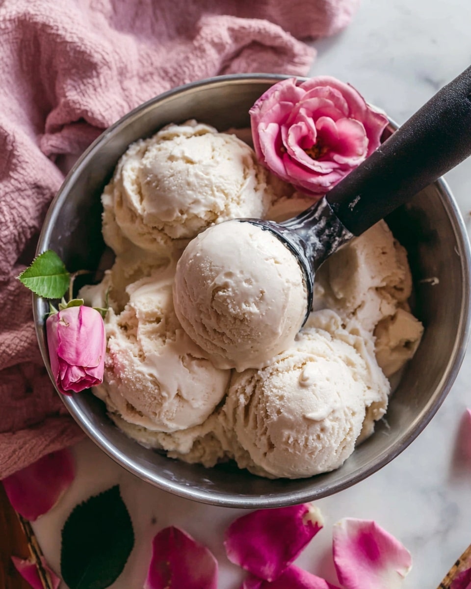 A metal bowl filled with several rough scoops of light beige ice cream, showing a creamy, smooth texture with small air pockets. A black ice cream scooper is inside the bowl, holding one rounded scoop of the same ice cream close to the top left side. On top of the ice cream, there is a single fresh pink rose with green leaves, partially resting on the scoops. Below the bowl, there are more pink rose petals scattered. The bowl sits on a white marbled surface with a soft pink cloth behind it. Photo taken with an iphone --ar 4:5 --v 7