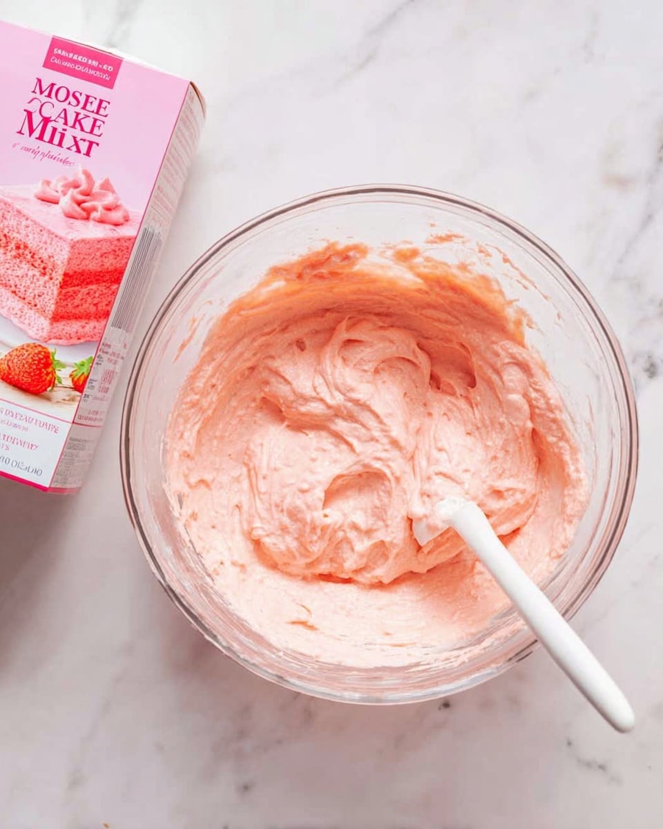 A clear glass bowl sits on a white marbled surface, filled with a smooth, pale pink cake batter that has a creamy, soft texture. A white spatula rests inside the bowl, partially covered in the same pink batter, showing it has been stirred. Next to the bowl is a pink box of cake mix with an image of a pink cake slice on it. The overall scene gives a fresh and clean look with a focus on the batter mixing process. photo taken with an iphone --ar 4:5 --v 7