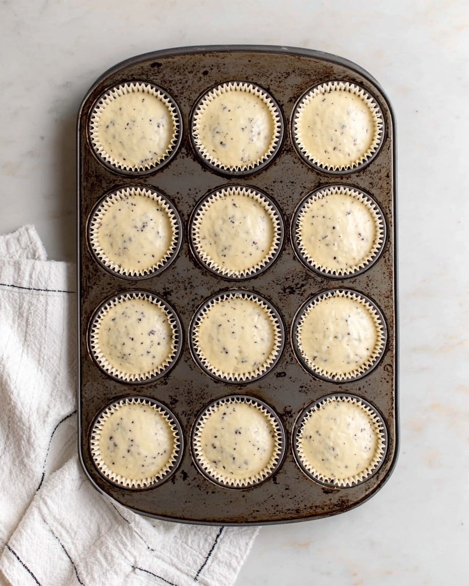 A dark metal muffin tray holds twelve cups, each lined with a light beige paper liner filled with creamy batter that has small dark specks and bits mixed evenly throughout. The batter fills each liner almost to the top, creating smooth, rounded surfaces. The tray is placed on a white marbled surface, and a white cloth with thin black lines peeks from the lower left corner. The lighting is soft and natural, highlighting the texture of the batter and the worn details of the tray. photo taken with an iphone --ar 4:5 --v 7