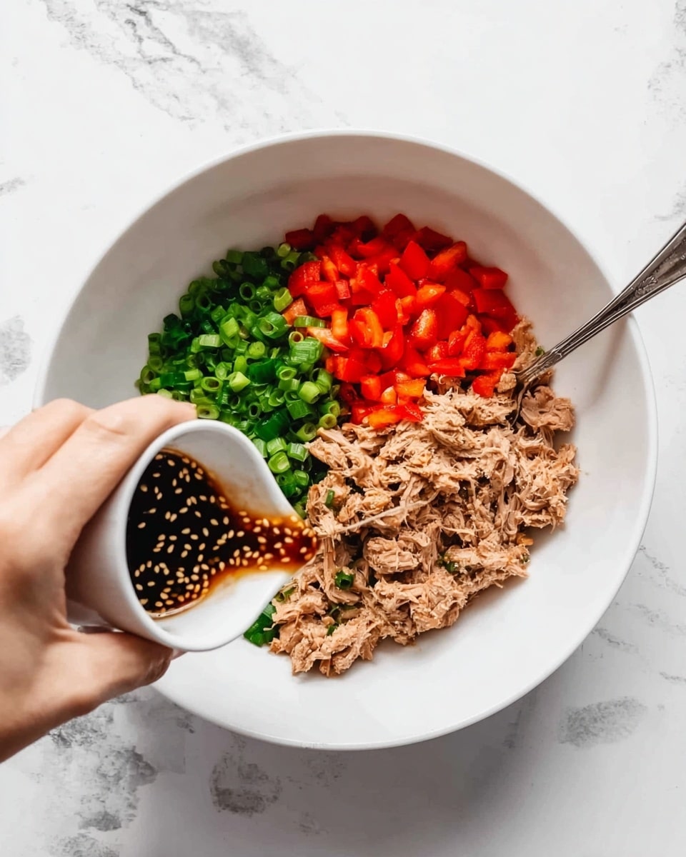 A white bowl sits on a white marbled surface, filled with three main layers: bright green chopped spring onions on the top left, vibrant red diced bell peppers below them, and light brown shredded tuna on the right. Dark brown sauce with sesame seeds is being poured from a small white cup held by a woman's hand from the left side of the image, adding texture and contrast over the tuna and vegetables. A silver spoon rests inside the bowl on the right edge, partially touching the tuna. Photo taken with an iphone --ar 4:5 --v 7