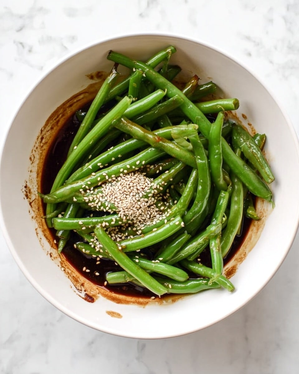 A white bowl filled with a layer of fresh, bright green green beans placed on top of a dark brown, thick sauce that is spread along the bottom and sides of the bowl. Some white sesame seeds are sprinkled over the green beans near the top left. The bowl is set on a white marbled surface. photo taken with an iphone --ar 4:5 --v 7