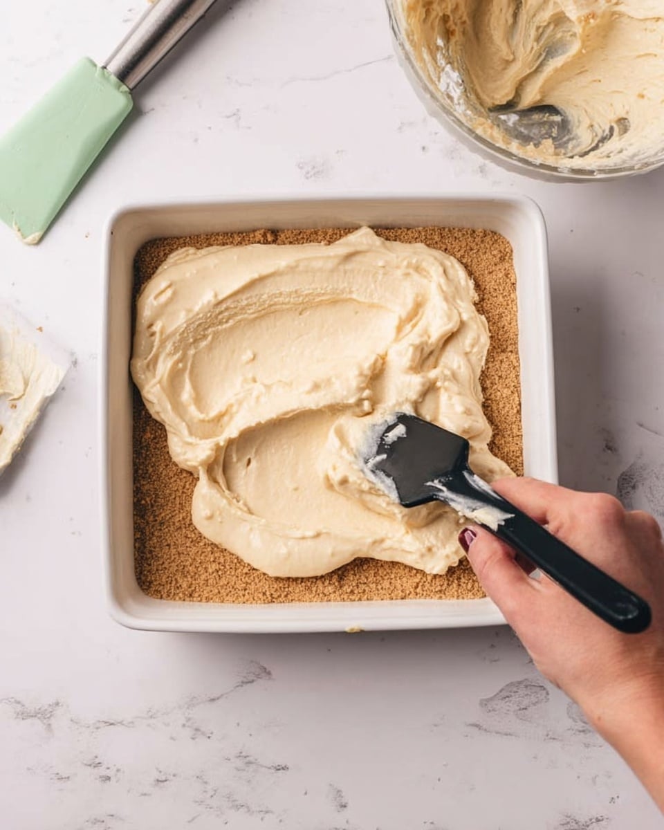 A white square dish with two visible layers inside is shown on a white marbled surface. The bottom layer is a smooth, brown crumb base spread evenly across the dish. The top layer is a thick, creamy light beige mixture being spread over the brown base by a woman's hand holding a black-handled spatula. To the right of the dish, a green spatula with some cream on it lies on the white marbled surface. Part of a bowl with some cream residue is visible in the upper right corner. Photo taken with an iphone --ar 4:5 --v 7
