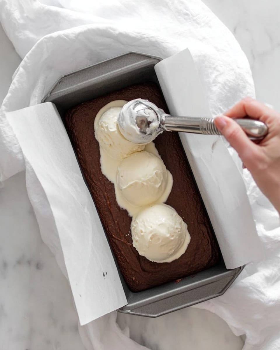 A rectangular gray baking pan lined with white parchment paper holds a thick dark brown chocolate batter spread evenly inside. On top of the batter are two large dollops of smooth white cream, placed close to each other near the center. A silver ice cream scoop held by a woman's hand is lowering a third dollop of the cream onto the batter. The pan rests on a white marbled surface with soft white fabric draped gently around it. photo taken with an iphone --ar 4:5 --v 7