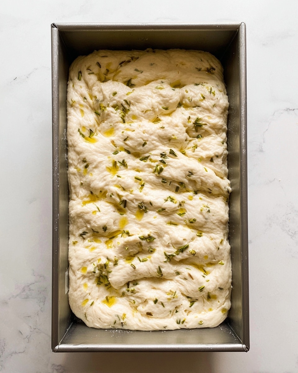 The image shows a rectangular metal baking pan filled with a thick, stretched dough that has a bumpy, cloud-like texture. The dough is pale cream color with some light yellow spots of oil glistening on top. Small green herb pieces are scattered evenly across the surface, adding specks of color. The pan sits on a white marbled surface, making the dough's colors and texture more visible. photo taken with an iphone --ar 4:5 --v 7
