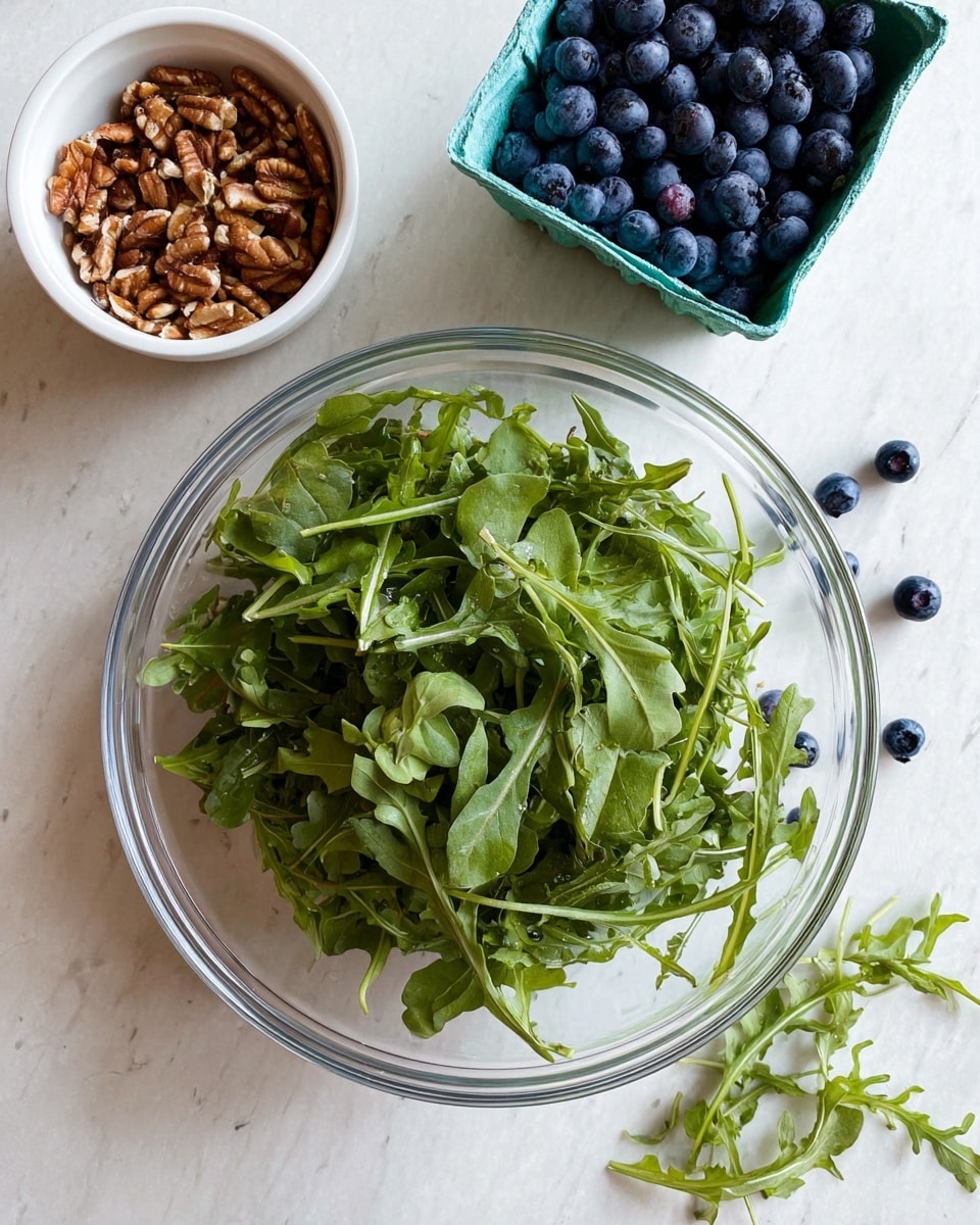 A clear glass bowl is filled with fresh green arugula leaves, showing a mix of smooth and slightly jagged textures, positioned at the bottom center of the image on a white marbled surface. Above the bowl, on the left, is a small white bowl filled with brown chopped nuts, adding a rough texture contrast. To the right of the nuts, there is a green cardboard container filled with dark blue, round blueberries, with a few scattered on the white marbled surface nearby. A few sprigs of arugula leaves are also lying loosely on the surface around the main bowl. photo taken with an iphone --ar 4:5 --v 7
