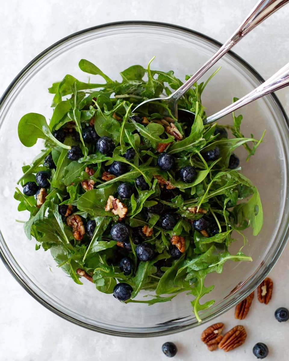 A clear glass bowl sits on a white marbled surface, filled with fresh green leaves of arugula that have a slightly textured, leafy appearance. Scattered throughout the arugula are round, shiny dark blueberries and small pieces of chopped pecans with a brown, crunchy look. Two silver forks rest inside the bowl at the edge, adding a metallic shine. A few blueberries and pecan pieces lie outside the bowl on the white marbled surface nearby. Photo taken with an iphone --ar 4:5 --v 7
