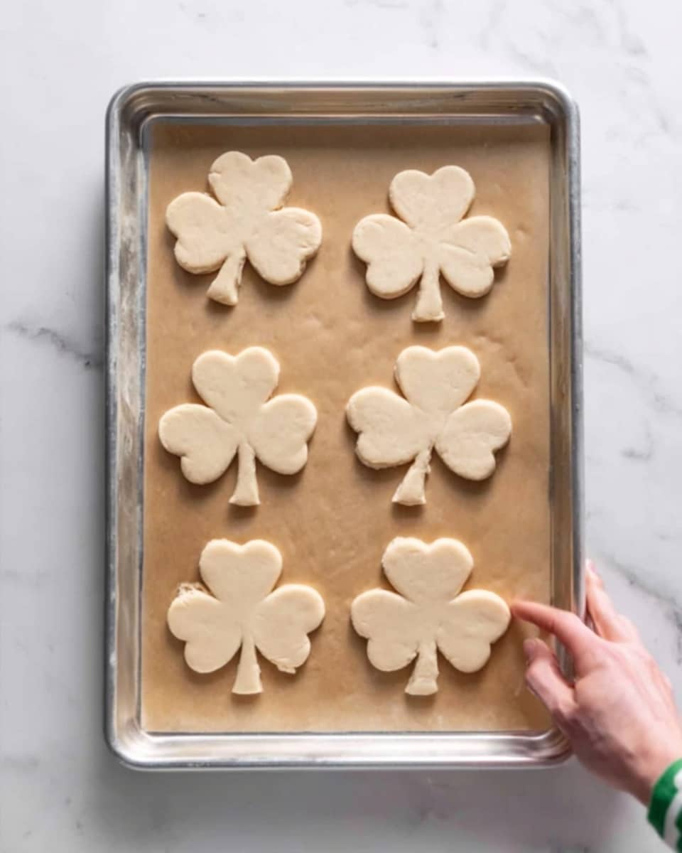 The image shows a silver baking tray on a white marbled surface with six raw dough pieces shaped like flowers evenly spaced on parchment paper. The dough is light beige and smooth with slightly thick edges, and a woman's hand is visible holding the right edge of the tray. Photo taken with an iphone --ar 4:5 --v 7