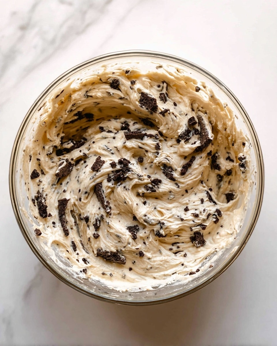 A clear glass bowl is filled with a creamy mixture that has a light beige color with dark brown chunks mixed throughout. The texture looks smooth but with many small bits giving it a speckled and slightly rough appearance. The mixture swirls inside the bowl, showing soft peaks and folds, and the bowl sits on a white marbled surface. Photo taken with an iphone --ar 4:5 --v 7