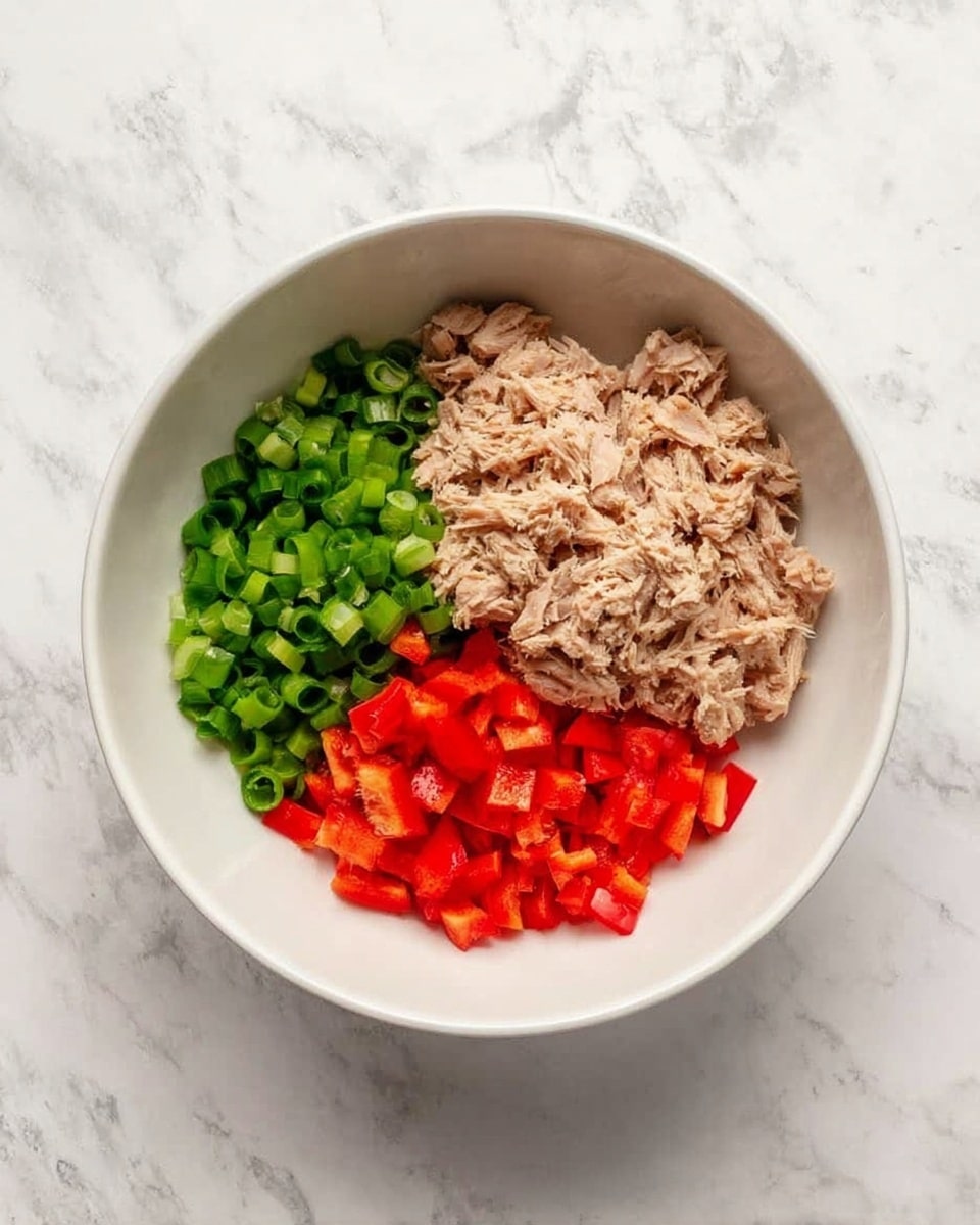 A white bowl sits on a white marbled surface, holding three distinct layers of ingredients arranged side by side. On the top right, there is a layer of light beige shredded tuna with a flaky texture. To the left of the tuna, a bright green layer of finely sliced green onions adds freshness. Below the green onions, chopped red bell peppers create a vibrant red layer, with small, evenly sized pieces. The bowl is clean and simple, highlighting the colorful ingredients inside. Photo taken with an iphone --ar 4:5 --v 7