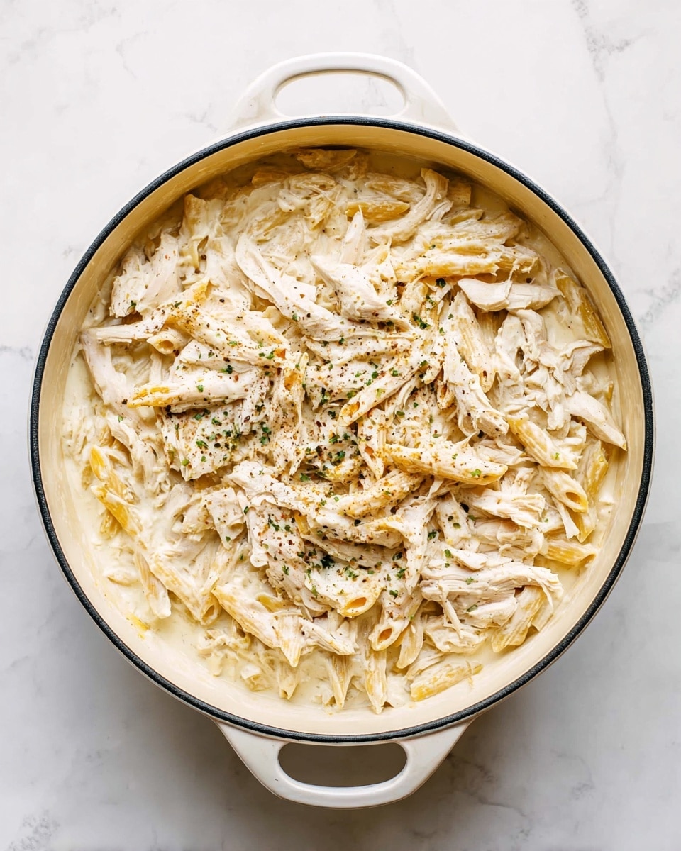 A white round pot with two handles holds creamy pasta mixed with shredded chicken. The dish has two main layers: the base is a thick white cream sauce and pasta pieces, mostly penne, with a soft texture. On top, shredded white chicken pieces are spread evenly with a light sprinkle of ground black pepper. The pot sits on a white marbled surface, adding a clean and bright look to the image. photo taken with an iphone --ar 4:5 --v 7